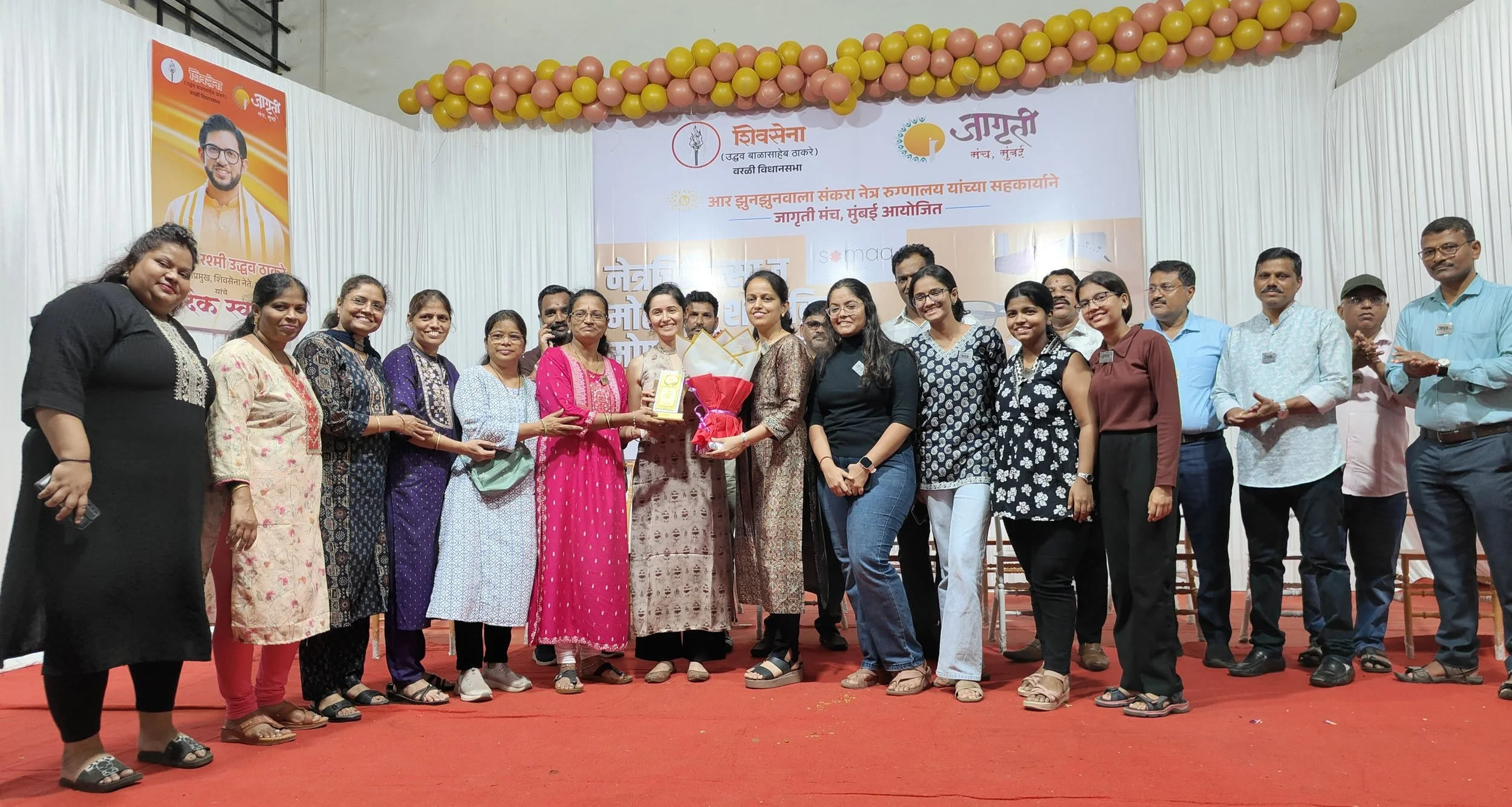 Group of people standing on a stage, with a woman in the center receiving a trophy and bouquet of flowers during an event or ceremony, decorated with balloons and banners.