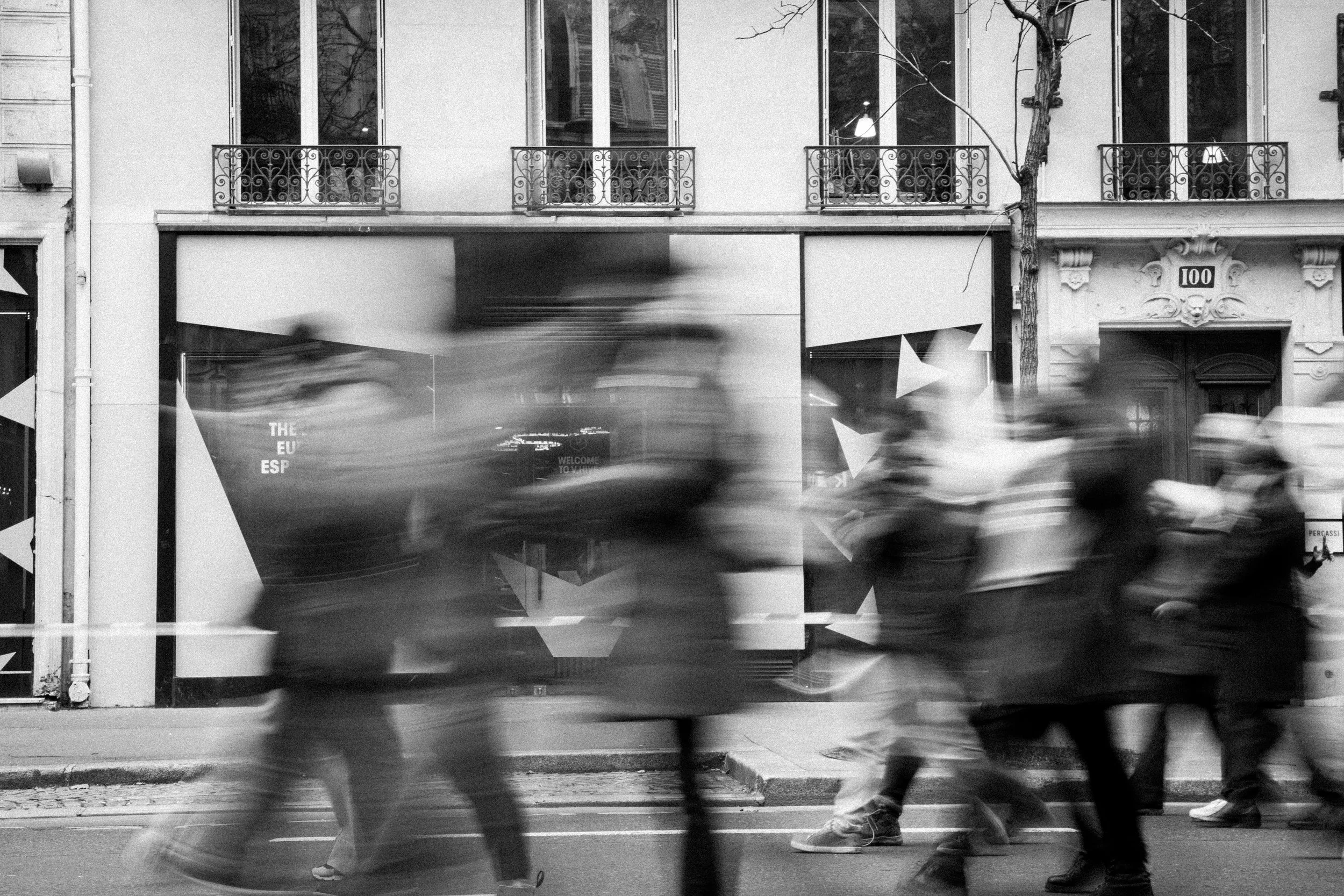 Blurry black-and-white photo of people walking quickly on a city street in front of a building with large windows and ornate iron railings.