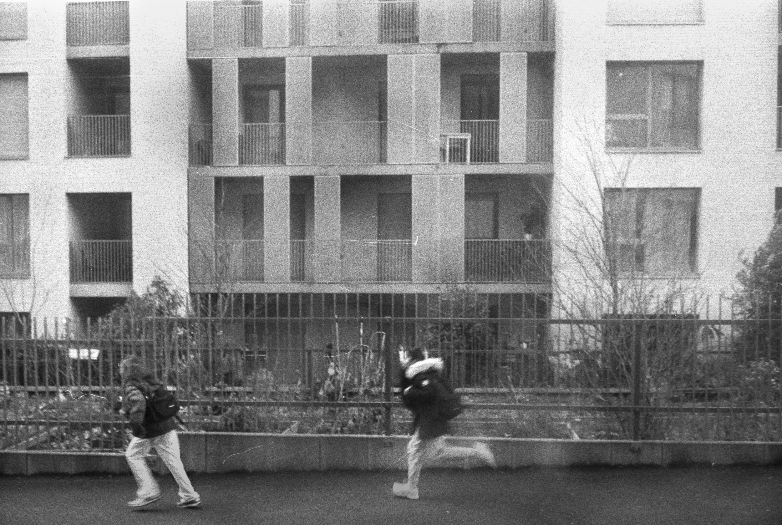 Black and white photo of two people walking along a sidewalk in front of a tall, modern apartment building with balconies. One person is wearing a backpack and a hoodie, while the other is in motion, possibly jogging, wearing a helmet and a jacket.