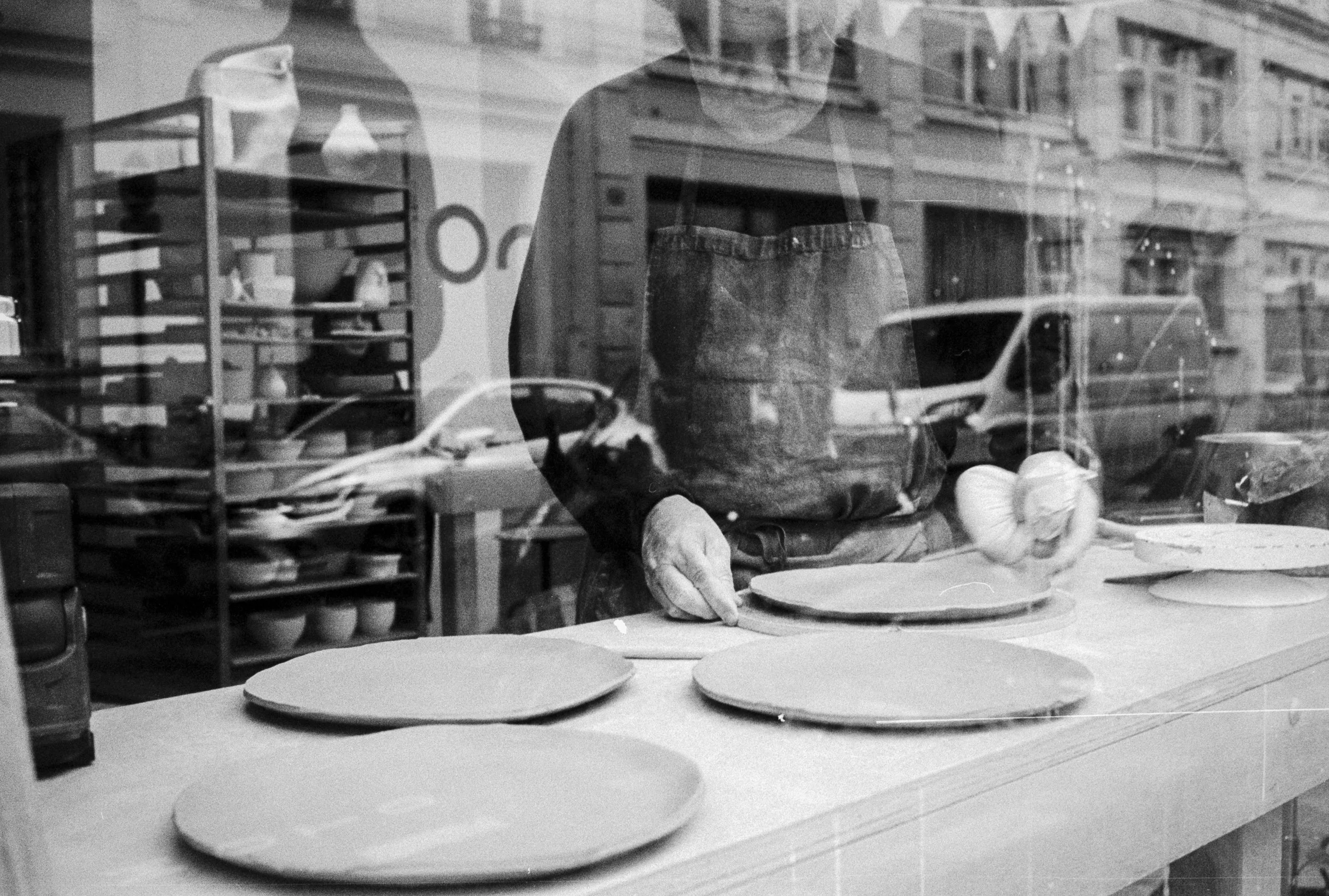 Black and white photo of a person working in a pottery studio, seen through a glass window, with shelves of pottery in the background and several clay plates on the table in front.