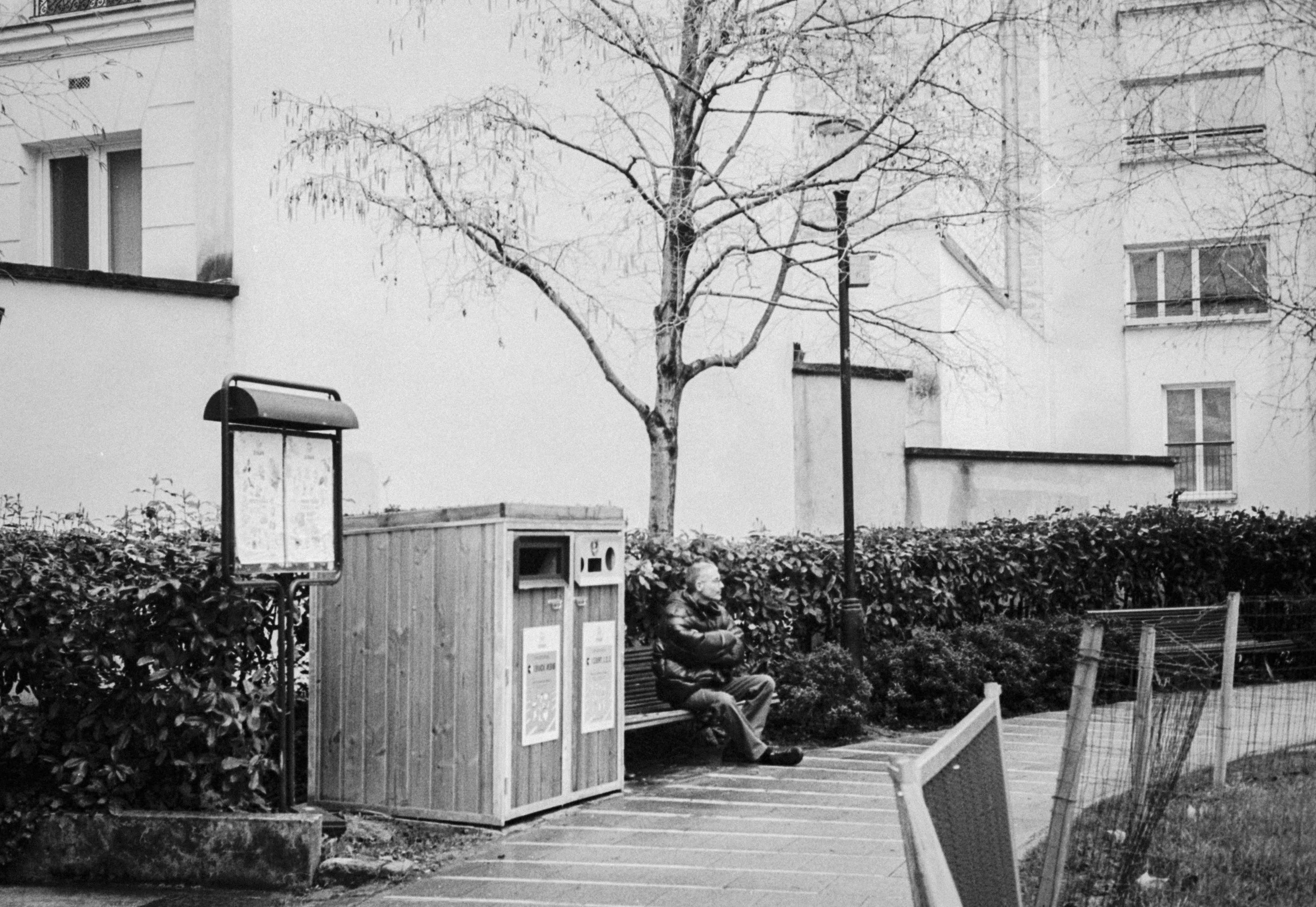 A man sitting on a park bench with his arms crossed in front of a tree and a hedge, near a public trash and recycling station, in an urban setting.