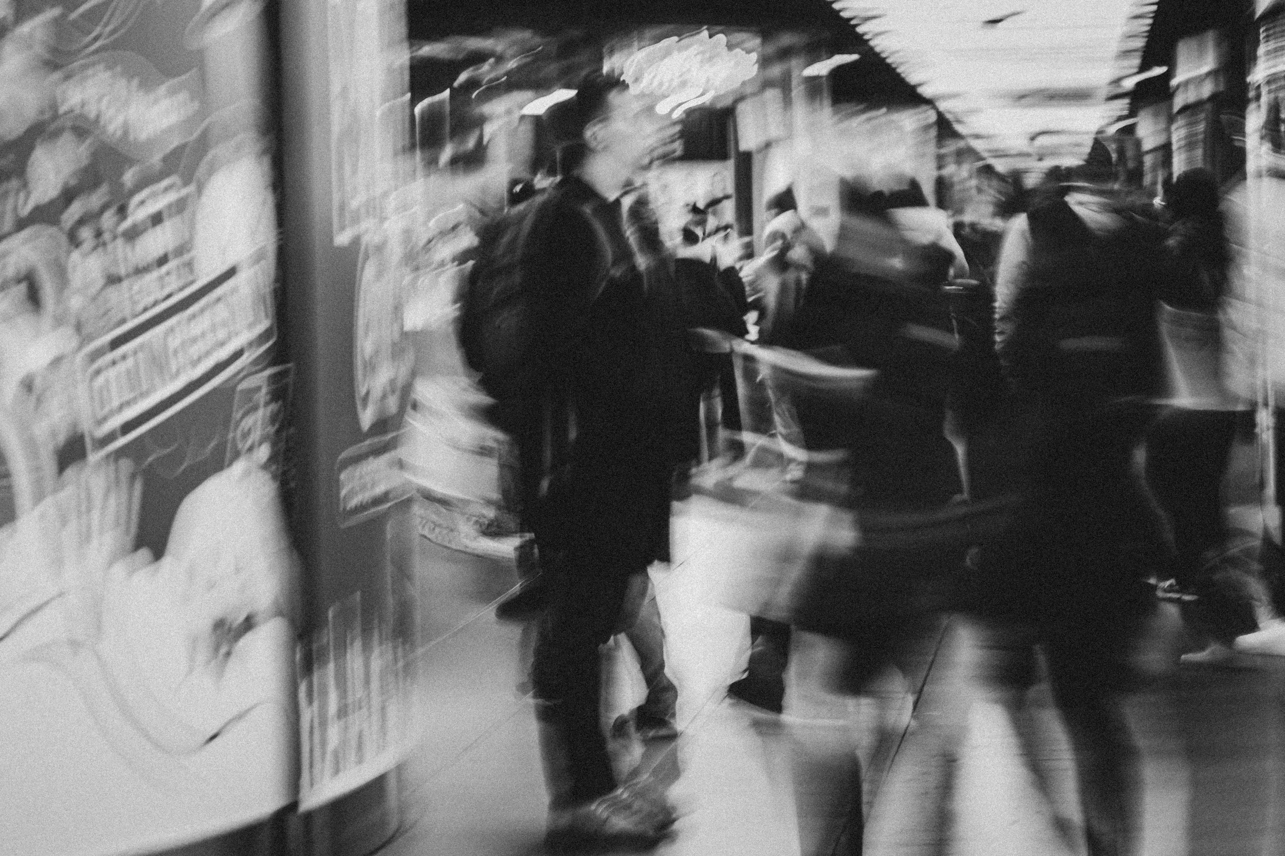 Blurry black and white photo of a hallway with people walking and standing, some holding items, and advertisements on the wall.
