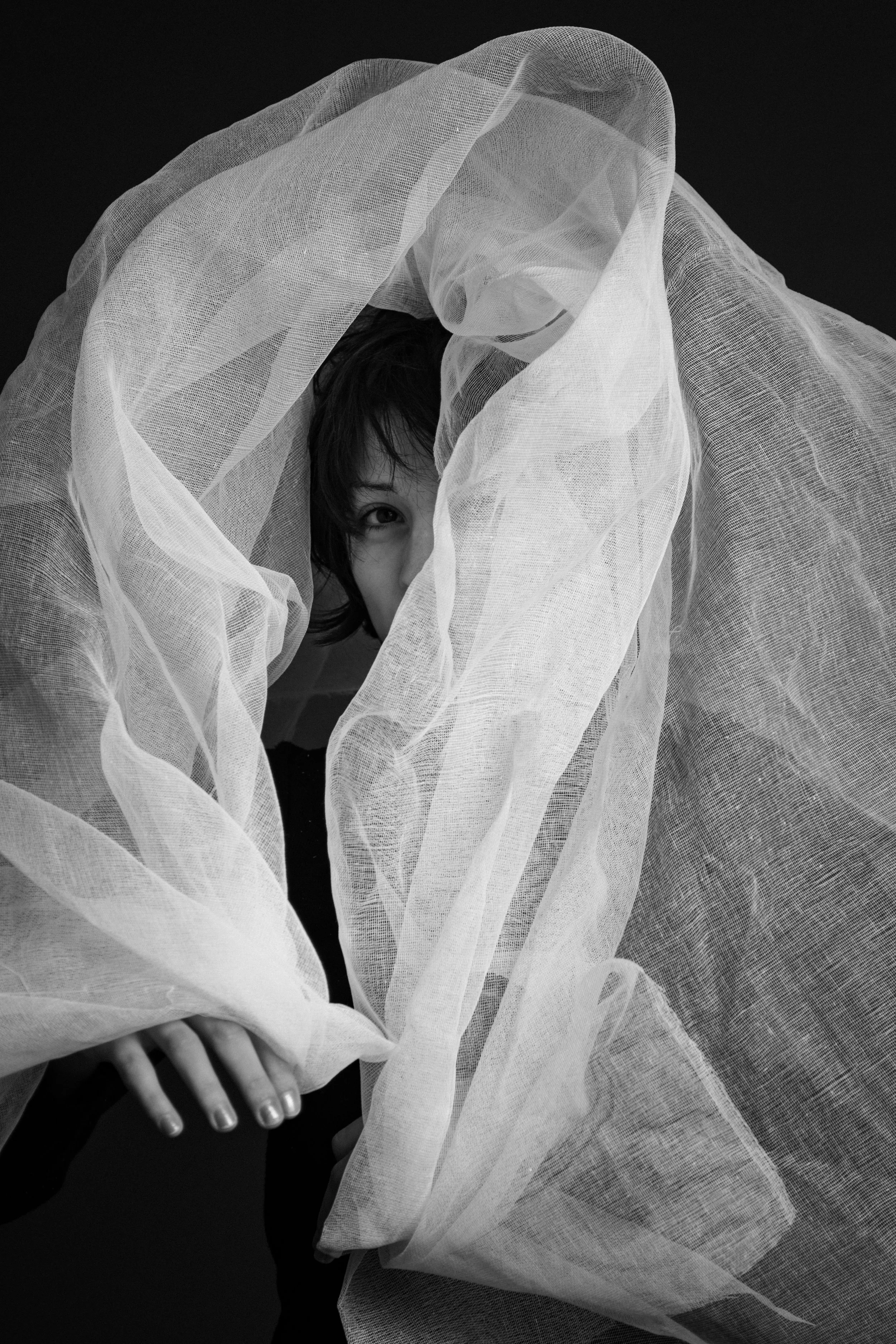 Person partially covered by sheer, light-colored fabric against a dark background, with one eye and hand visible.