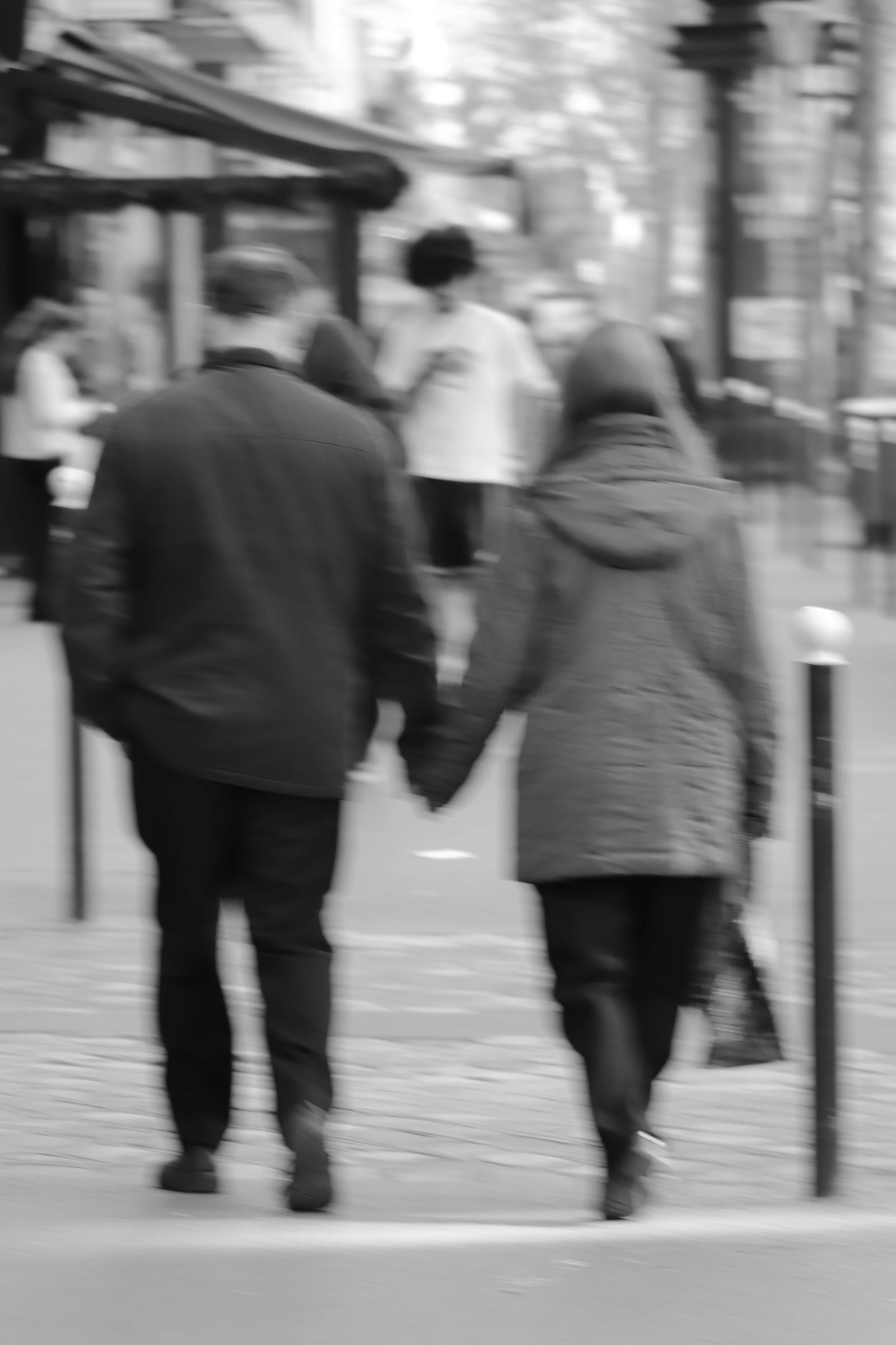 A black-and-white photo of a couple holding hands walking on a city sidewalk, with people and buildings in the background.