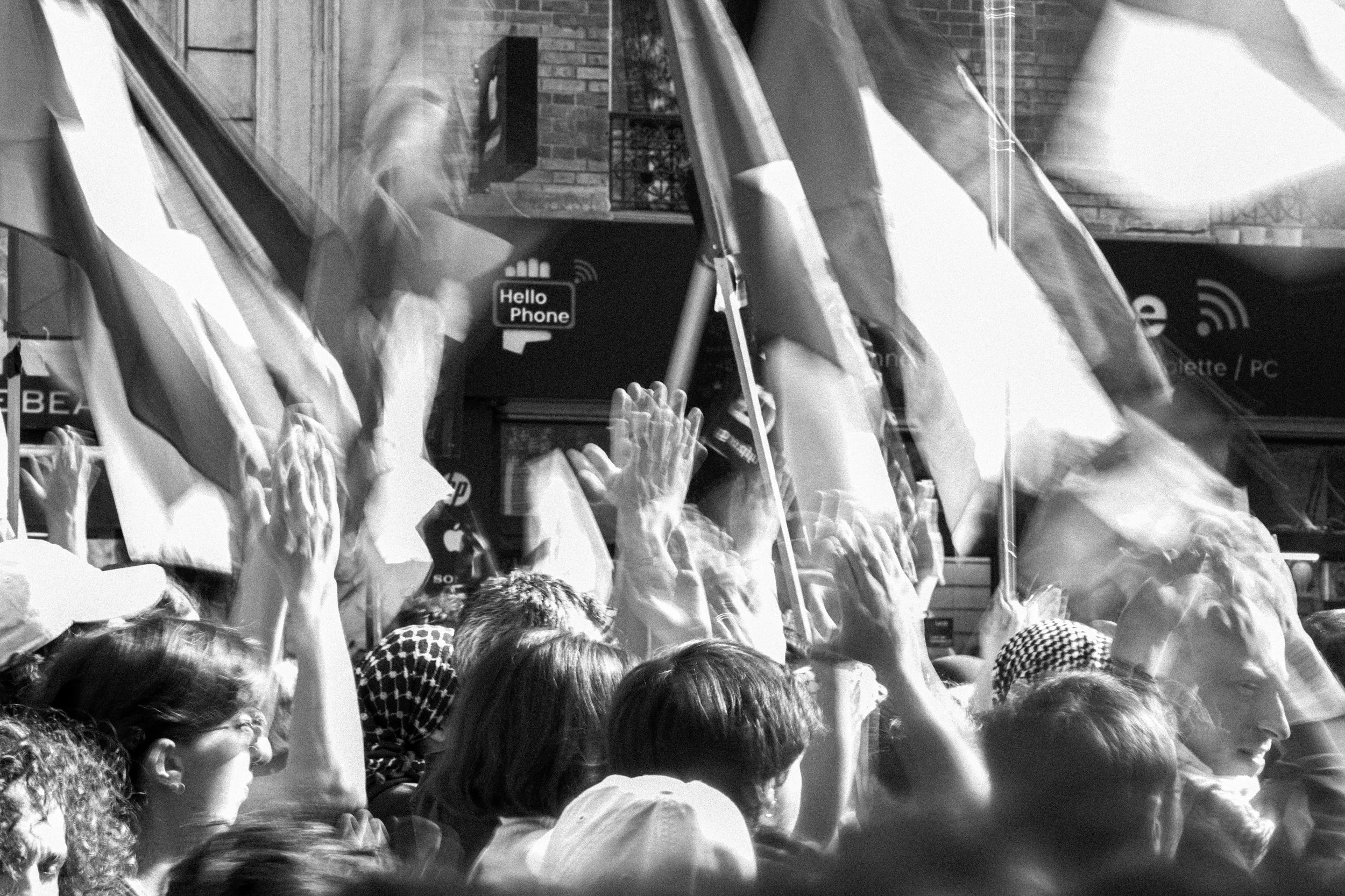A black and white photo of a crowd of people raising their hands in a public gathering, with some holding flags or banners. The faces and details are obscured by motion blur.