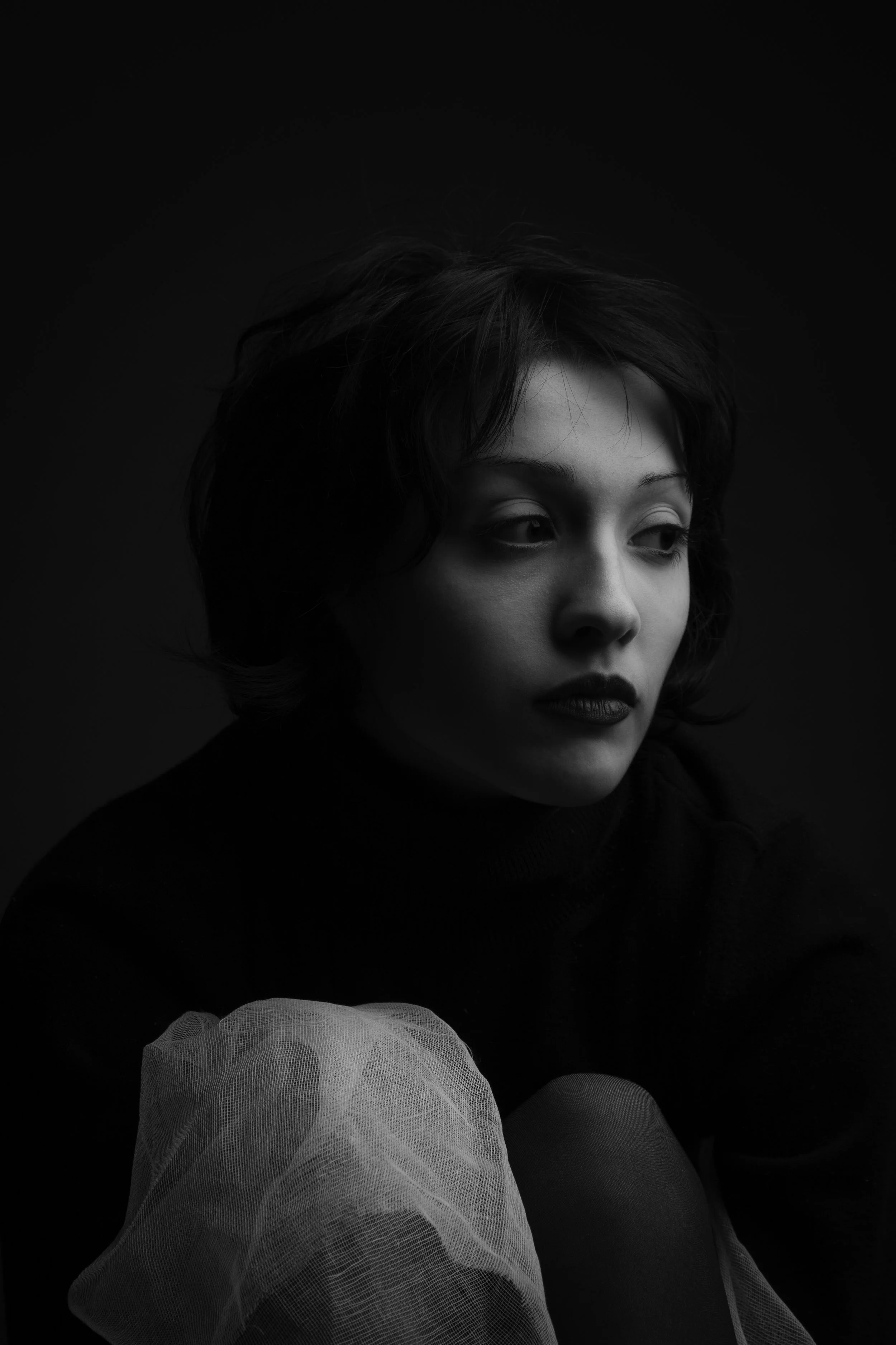 A black and white portrait of a woman with short dark hair, wearing dark lipstick, a dark top, and a light-colored tulle fabric in the foreground.