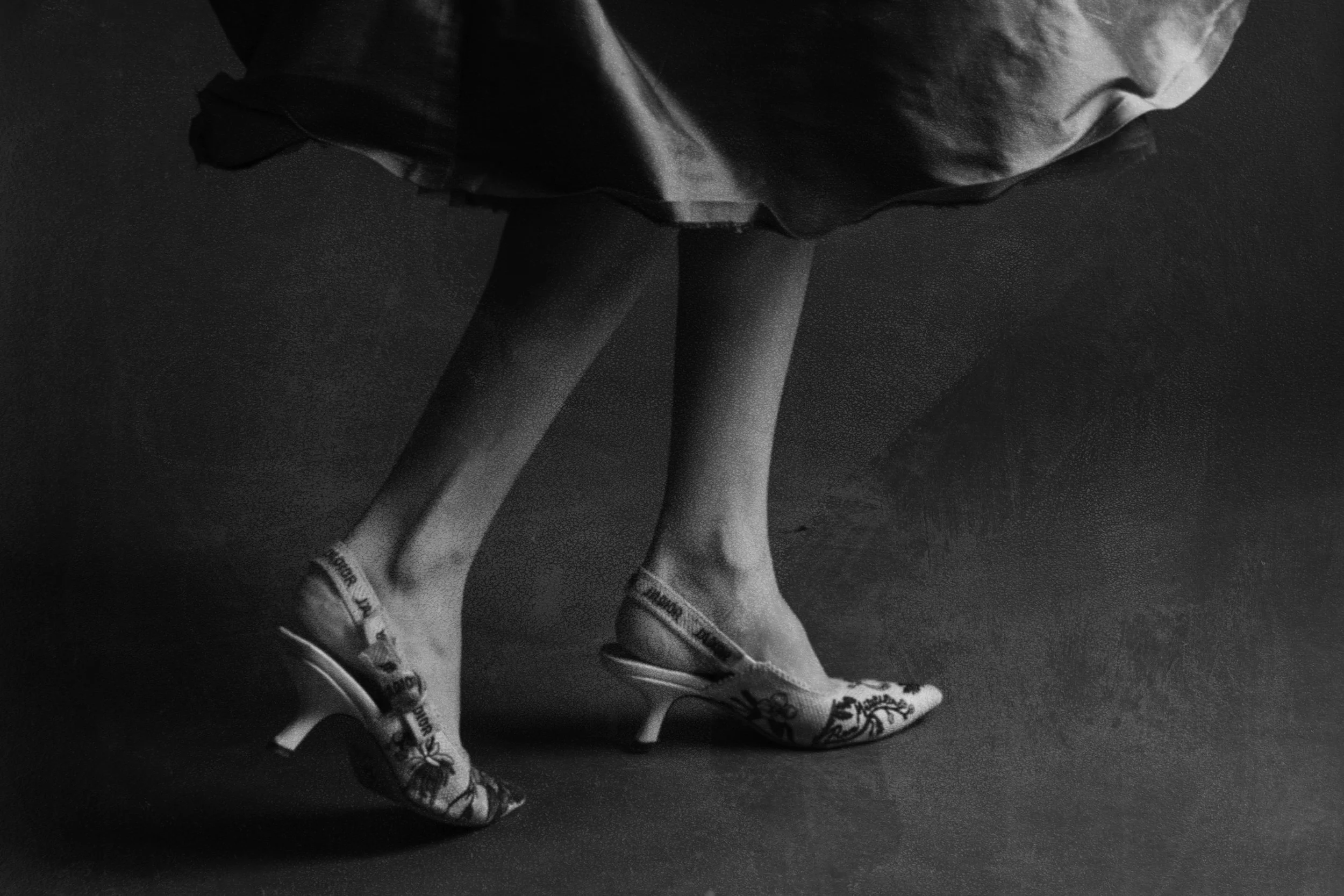 Black and white photo of a woman in a skirt and patterned high-heeled shoes, with only her legs visible, standing on a plain surface.