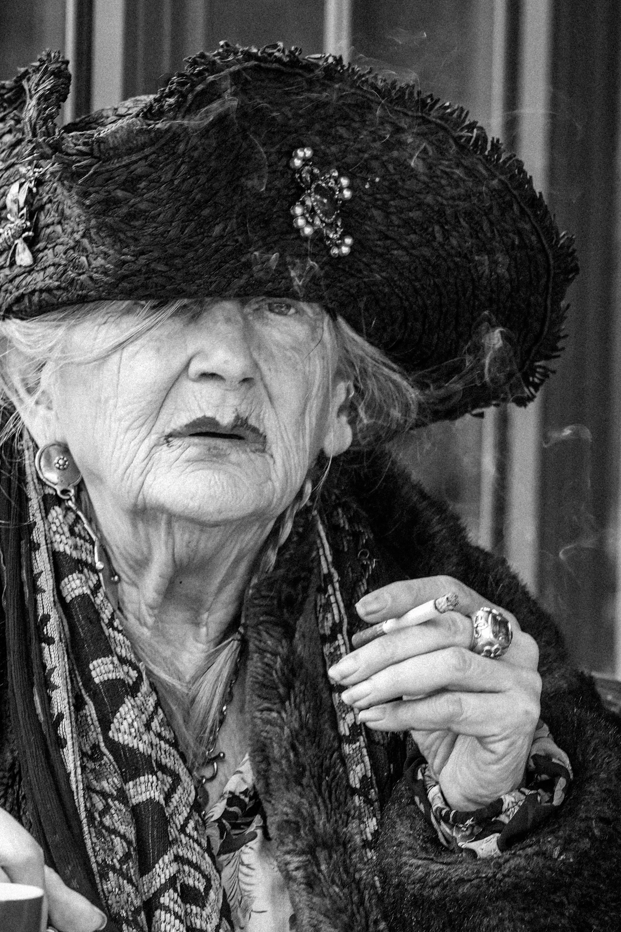 An elderly woman wearing a large, decorated hat, earrings, and necklaces, smoking a cigarette, with a contemplative expression.