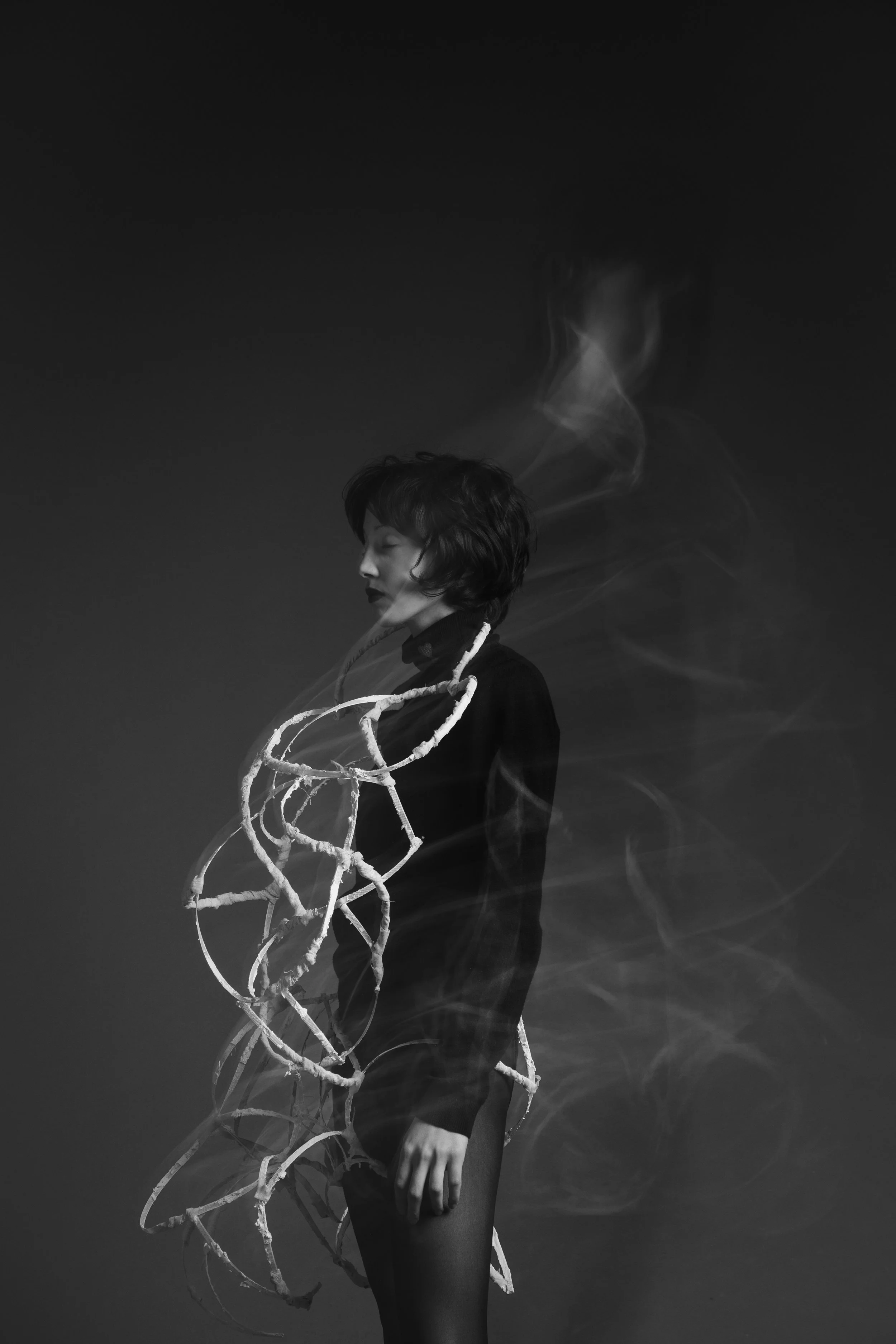 A black and white photo of a woman with short hair standing in profile. She is wearing a black outfit with a sculptural, abstract, rope-like feature around her shoulders and chest. Smoke swirls around her, adding a surreal, artistic effect.