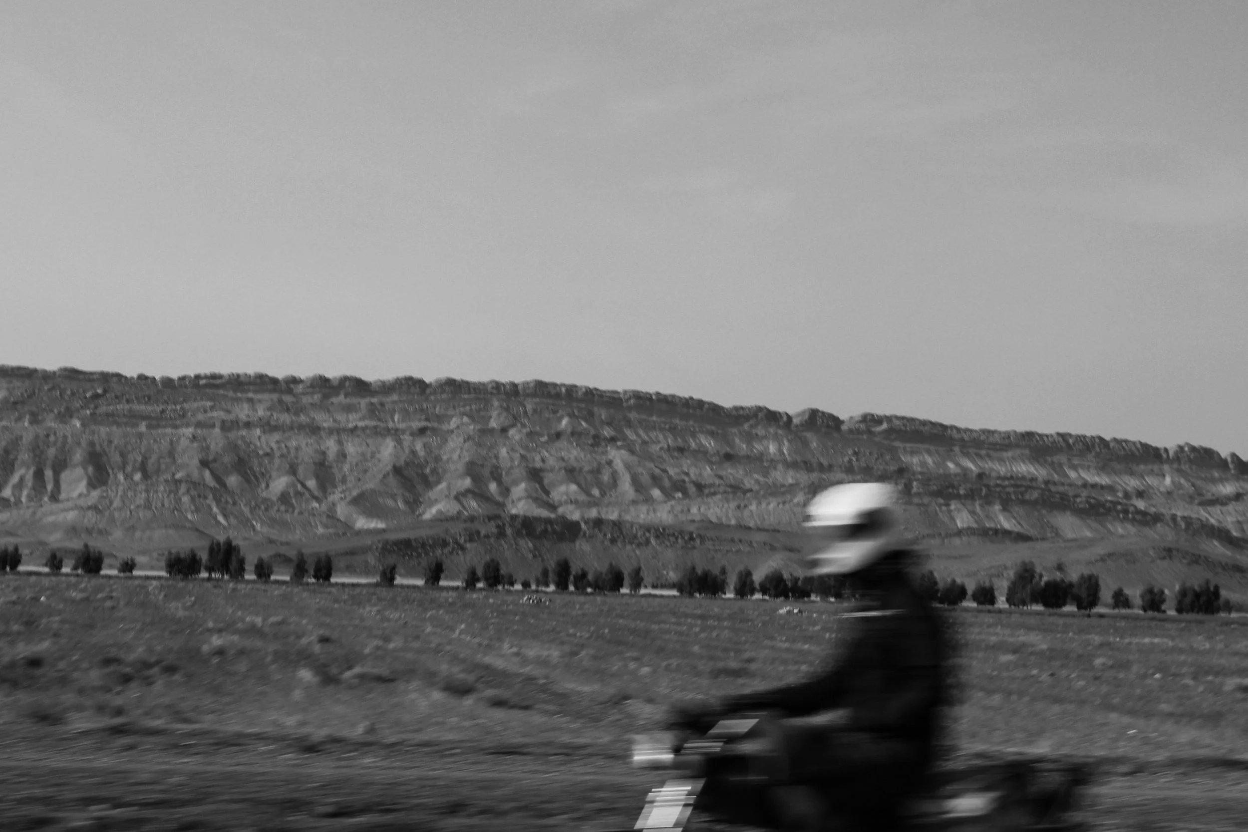 A person wearing a helmet riding a motorcycle or scooter on a road in a rural area with a hillside and trees in the background, in black and white.