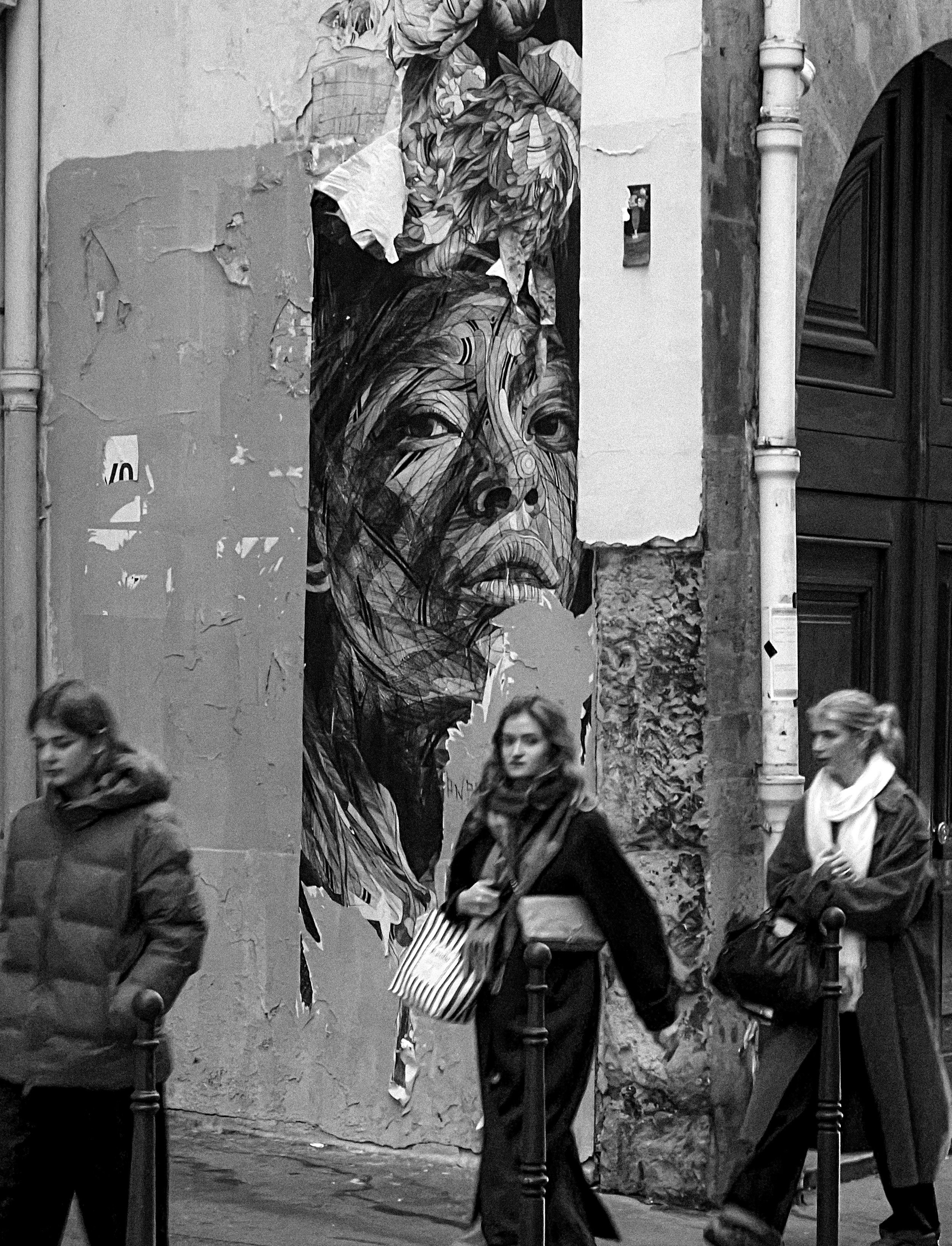 Black and white photograph of street scene with three women walking past a large mural of a woman's face on a worn wall. The mural depicts a woman with long hair and a serious expression.