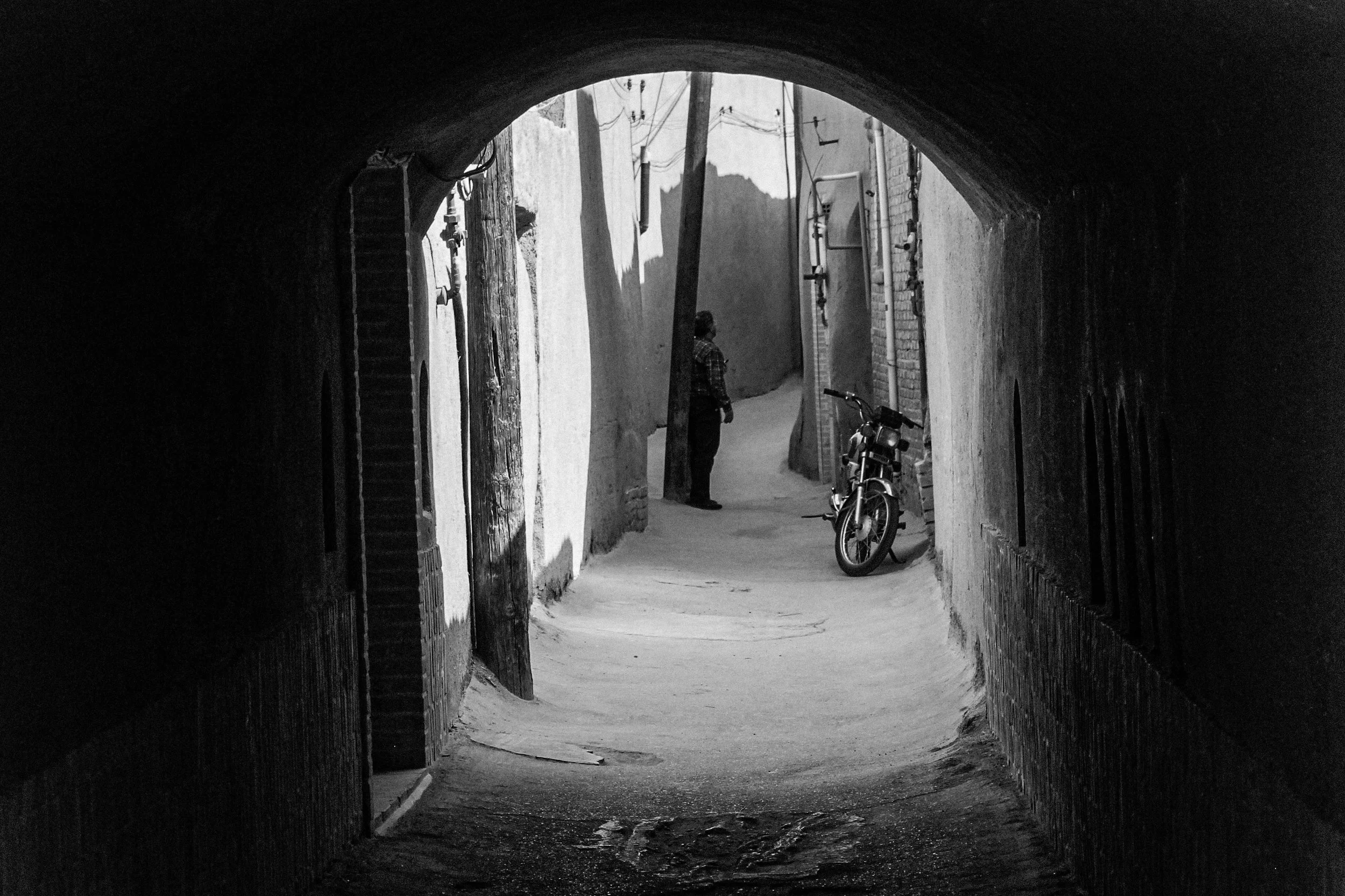 A black and white photo of a narrow alleyway viewed from inside a tunnel, with a person standing at the far end near a parked motorcycle.