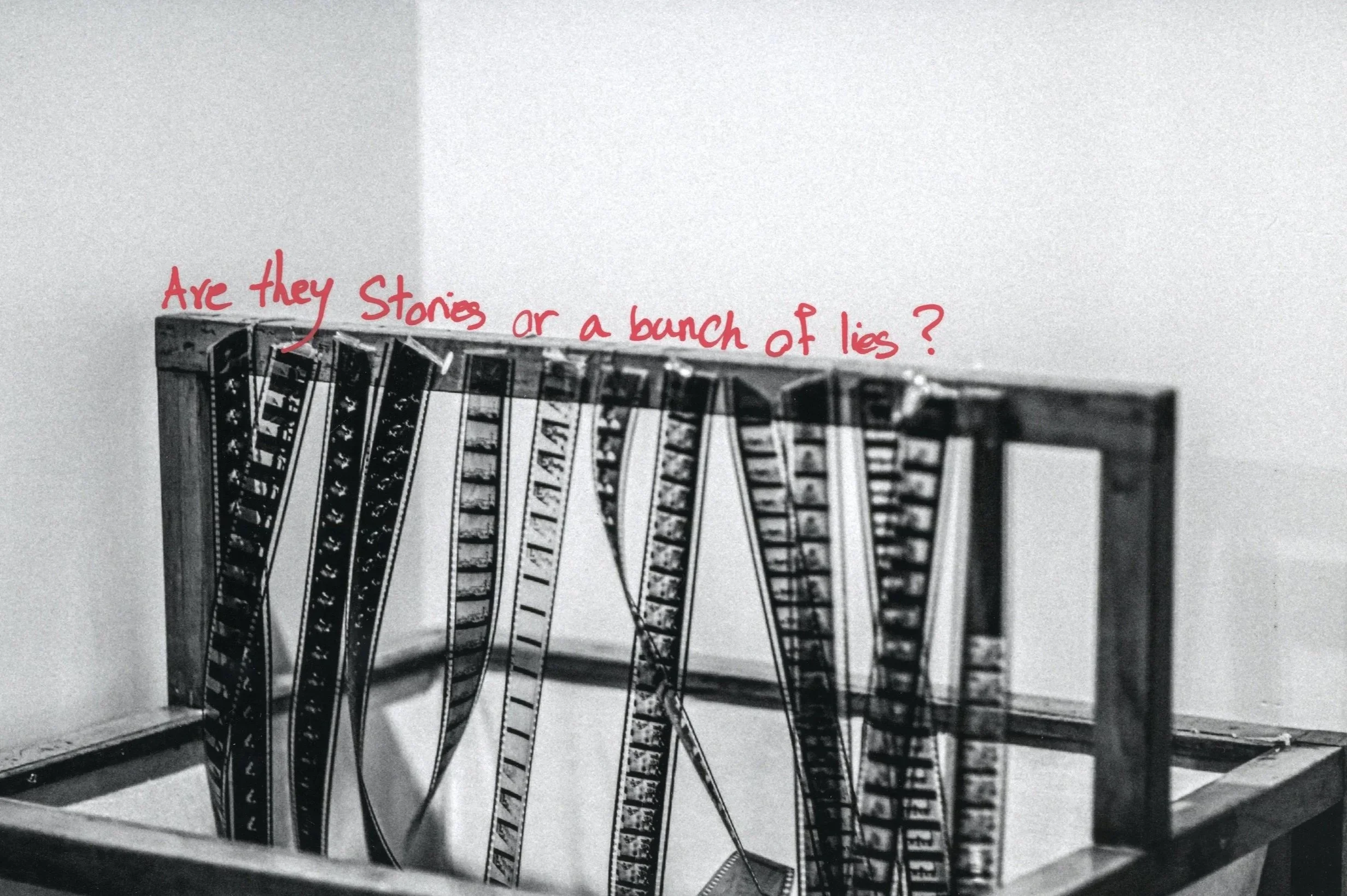 A partially built wooden picture frame with strips of film hanging inside, and red handwritten text on the wall reading 'Are they Storie or a bunch of lies?'