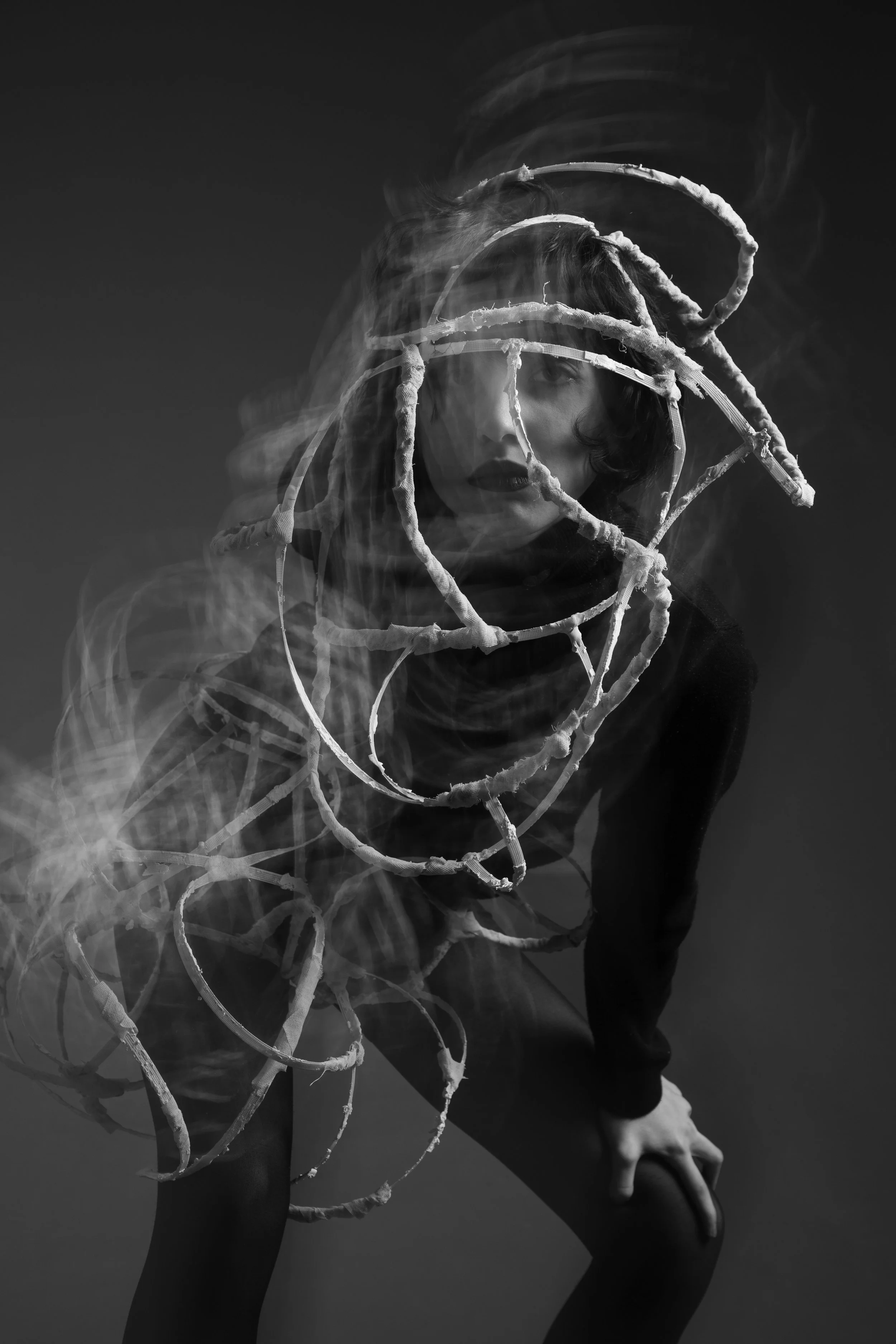 Black and white photo of a woman with short hair and dark lipstick, surrounded by a spiral of skips and smoke, leaning forward with one knee bent and hand on her thigh.