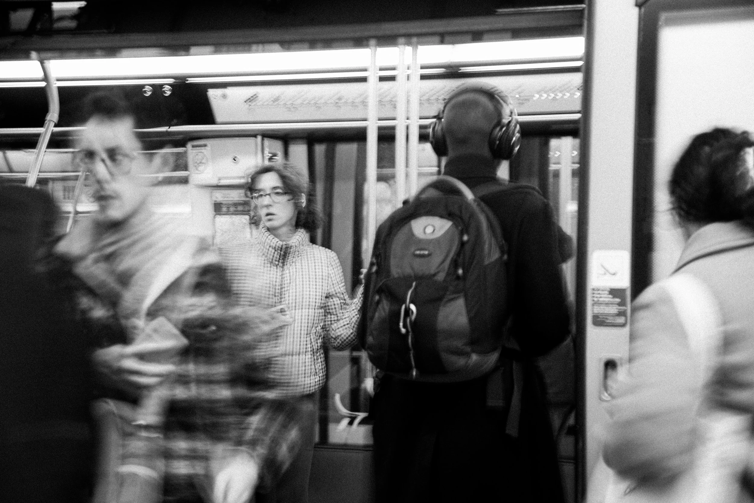 Blurred image of people standing and waiting on a subway train platform. One woman with glasses in a checkered shirt looks surprised or confused, and a man with a backpack and headphones is facing away. Other people are partially visible.