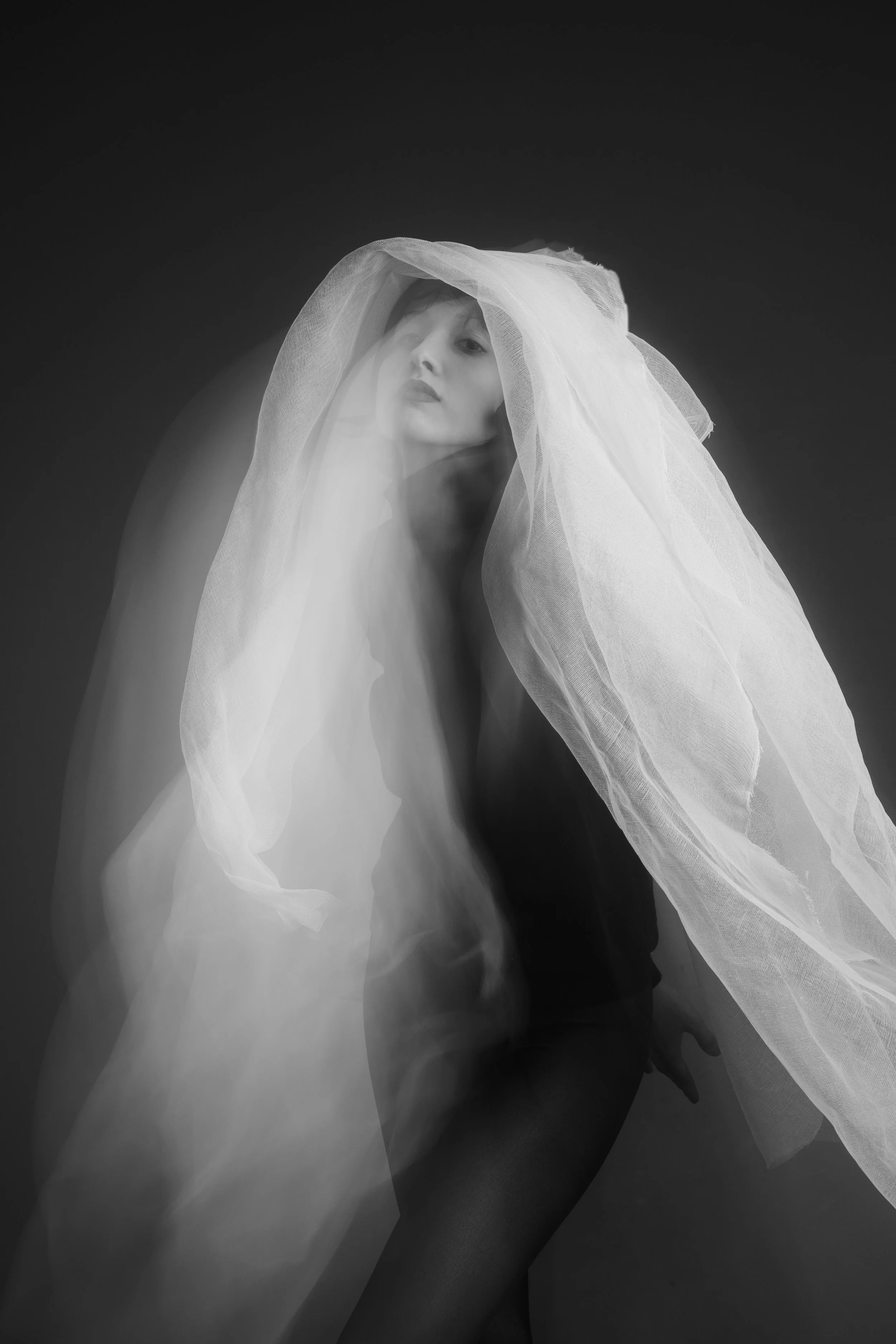 A woman with a veil over her head, posing against a dark background in black and white, with a ghostly, blurred motion effect.