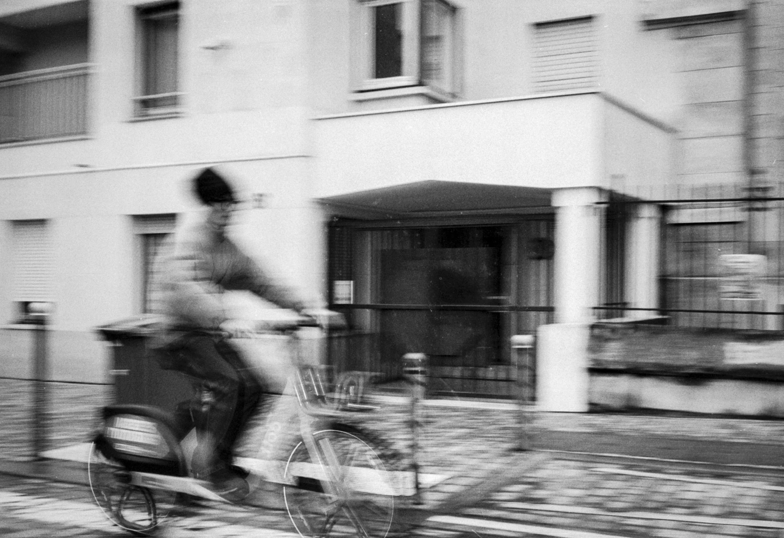 Black and white photograph of a person riding a bicycle near a modern apartment building with balconies and large windows.