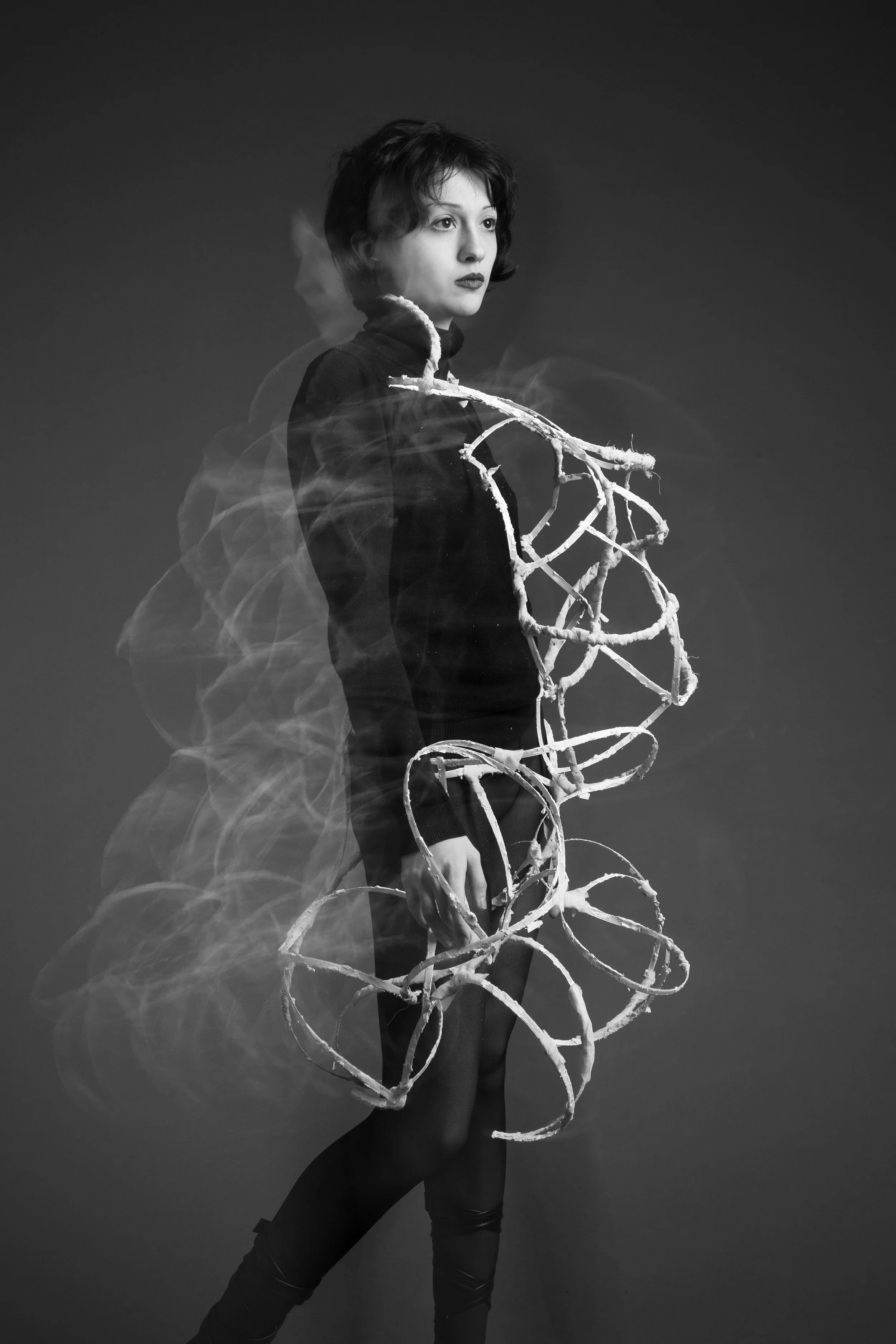 Black and white photo of a woman with short dark hair, dressed in dark clothing, standing against a plain background, with a spiral wire sculpture around her body.