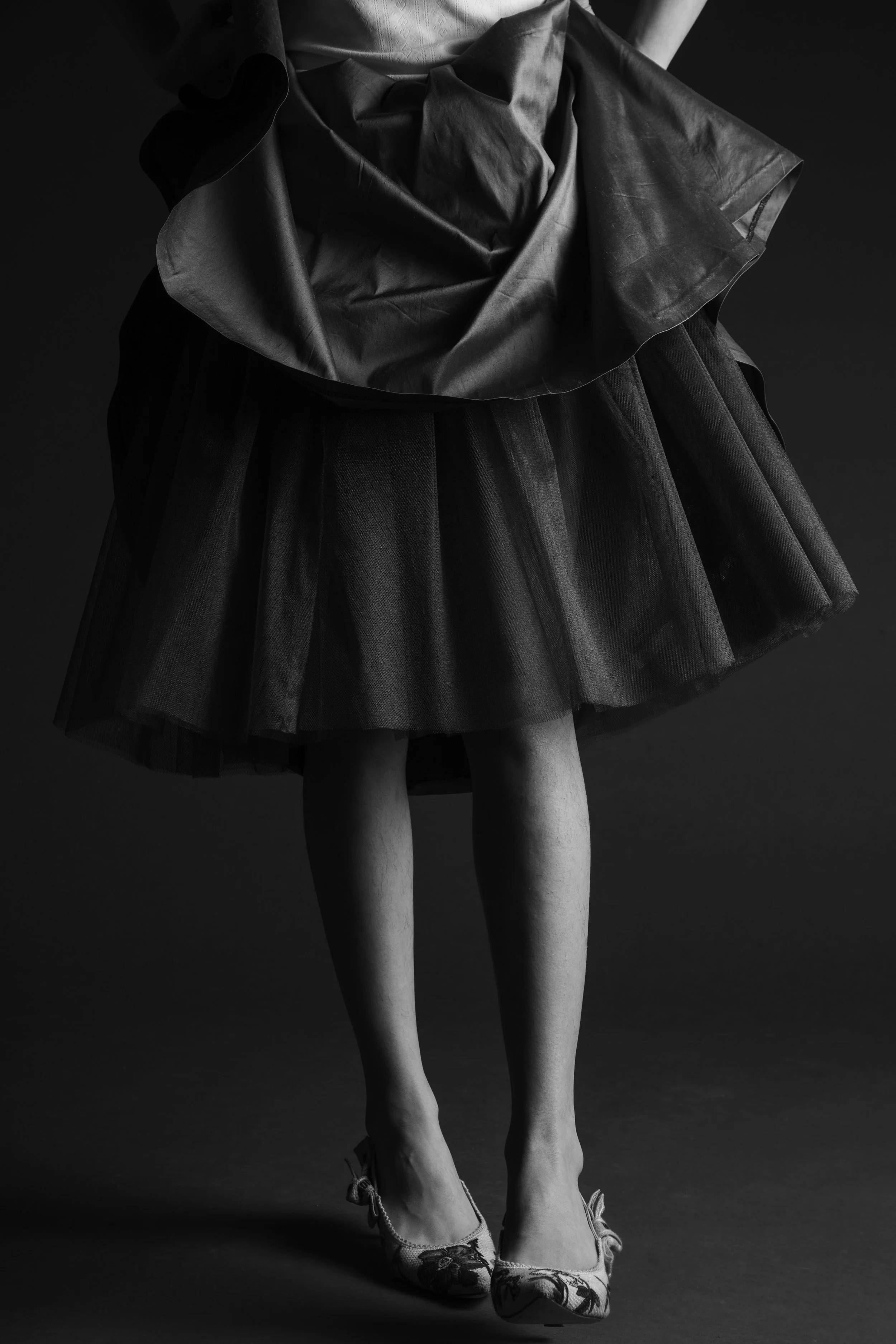 A black and white photograph of a person wearing a dark tulle skirt, a shiny top, and floral high heels, with their legs visible from the knees down.