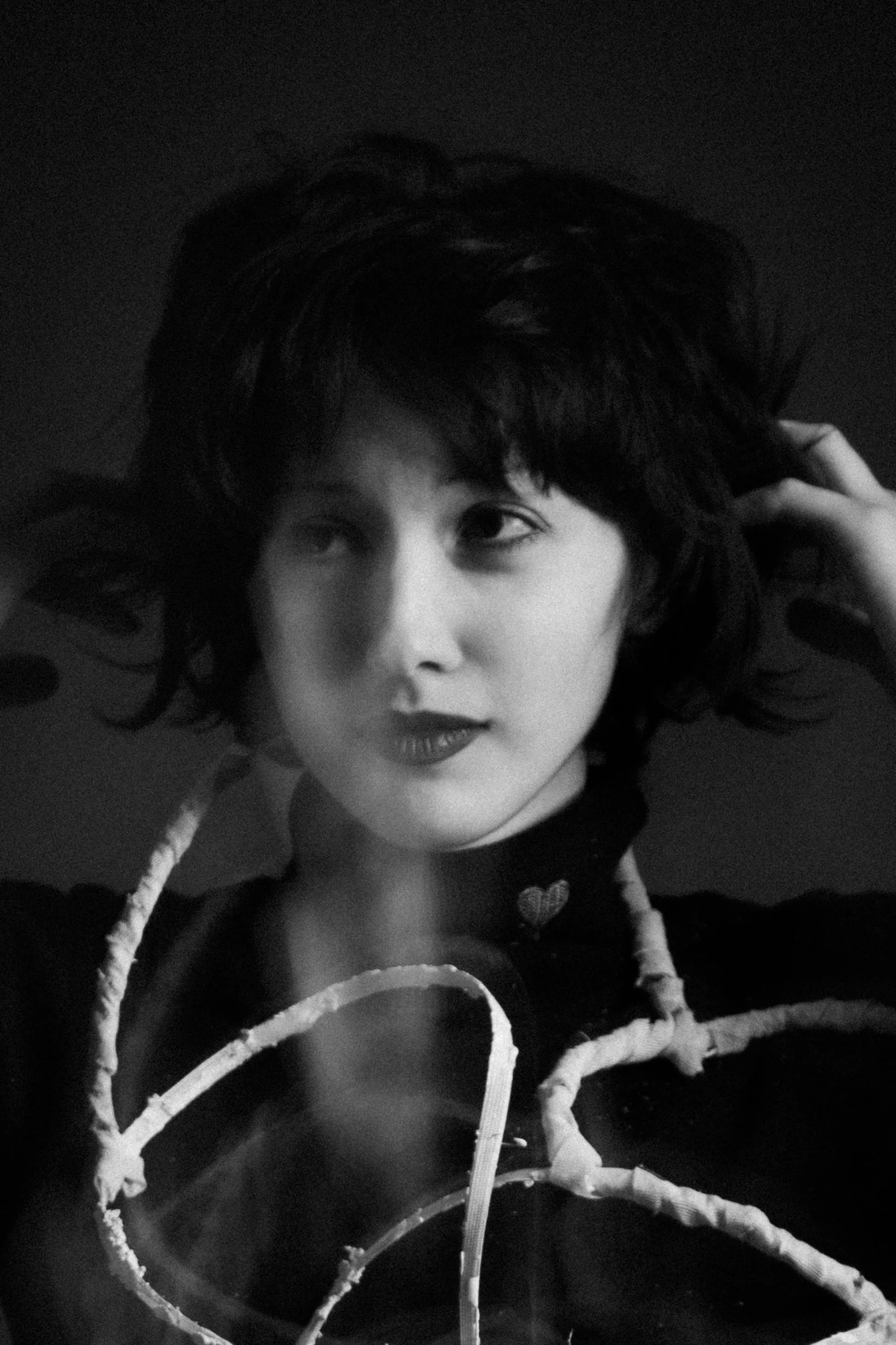 A black and white portrait of a young woman with short, curly hair. She is touching her hair with one hand, looking slightly to her right. She is wearing a dark top with a heart-shaped detail on the collar and large hoop earrings.