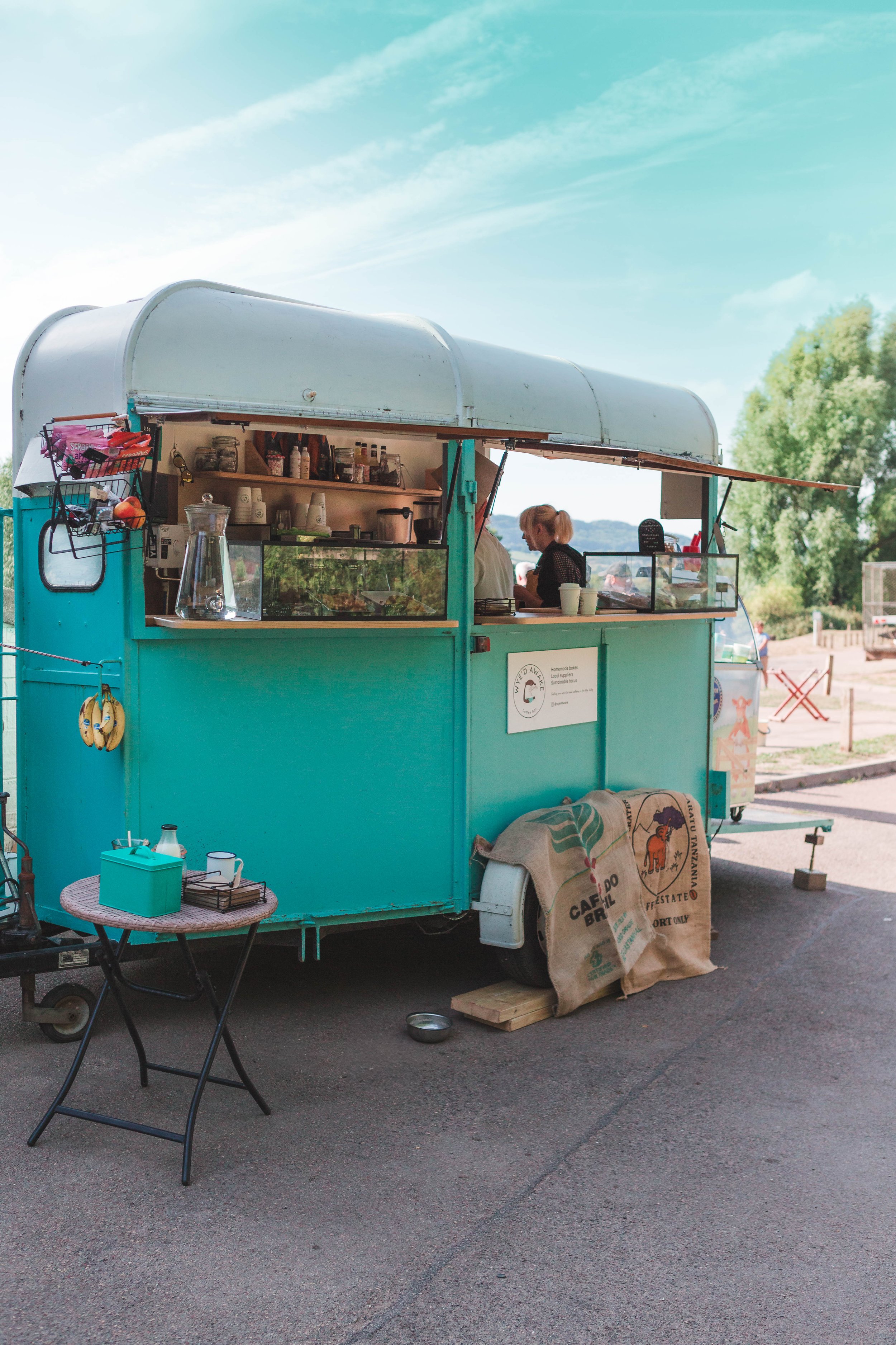 A teal food truck with an open window serving customers outdoors on a clear day. The truck displays various bottles and jars, and a woman is seen inside. There is a small round table with a teal box, a clipboard, and a black chair in front of the truck. A sack of coffee beans and a metal bowl rest on the ground. The background includes green trees and a blue sky with some clouds.