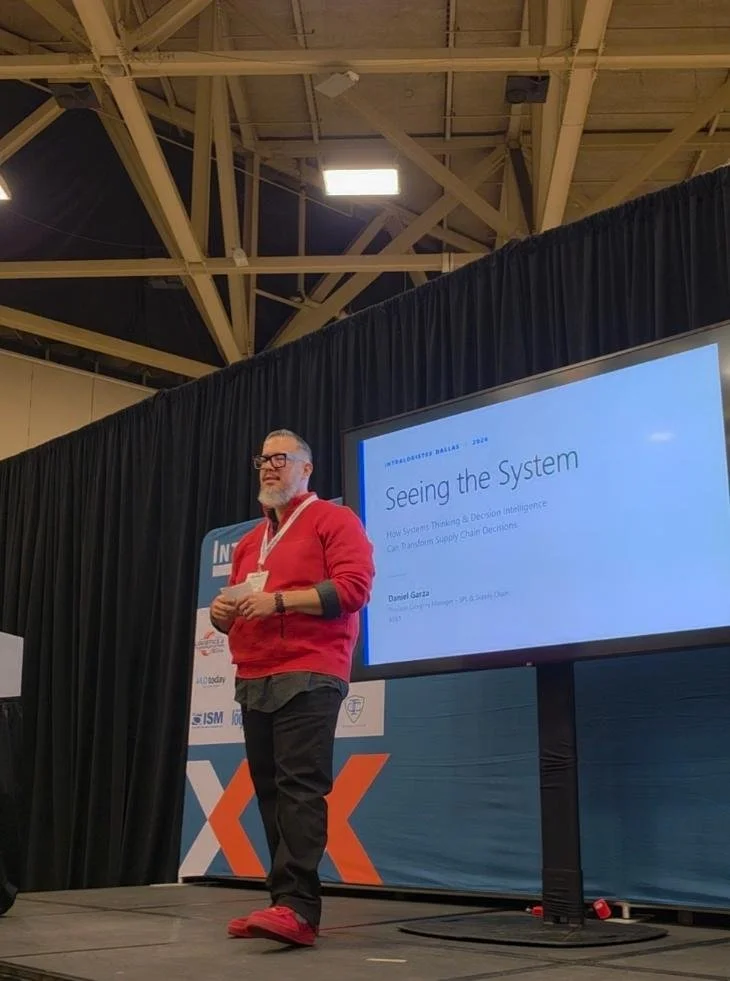 A man in a red jacket and dark pants giving a presentation at a conference, standing on a stage with a large screen displaying a slide titled "Seeing the System".