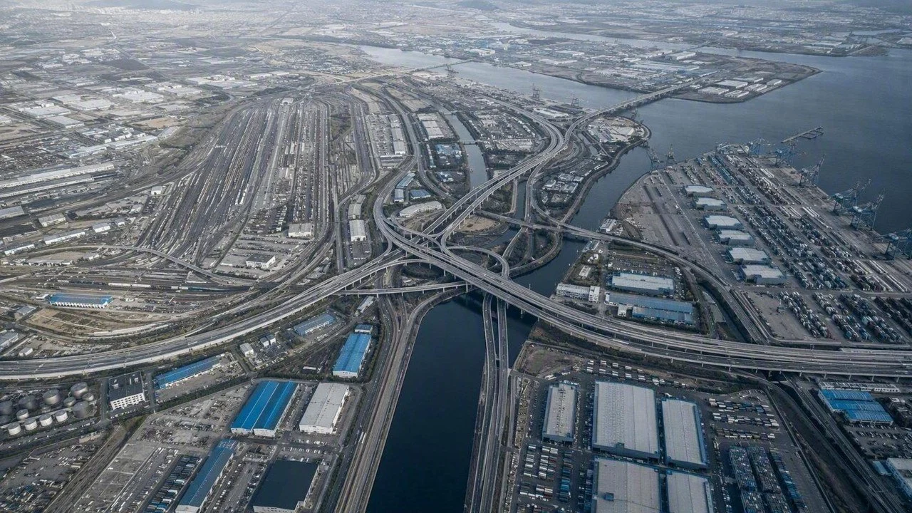An aerial view of a complex highway interchange over a body of water in an industrial area, with shipping docks and cranes visible along the waterfront.