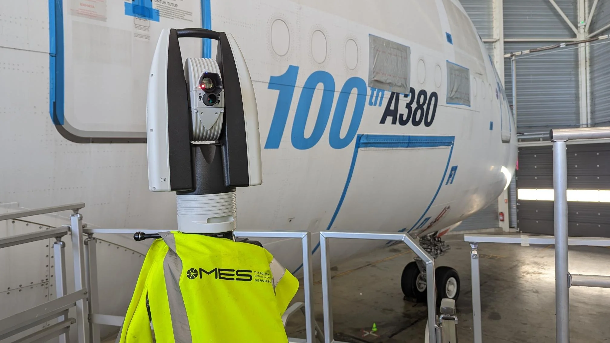 Close-up of a ground-scanning radar device positioned in front of a large airplane in a hangar. The airplane has blue and black text on its fuselage that reads '100 TH A380.' A high-visibility yellow jacket with the logo 'MES' is draped over a platform in front of the radar device. 