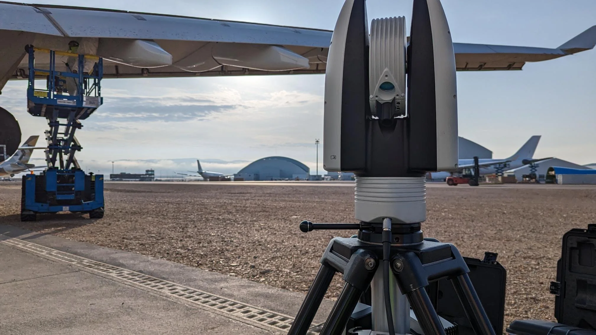A radar or laser scanning device on a tripod positioned on an airport tarmac, with airplanes and airport hangars in the background.