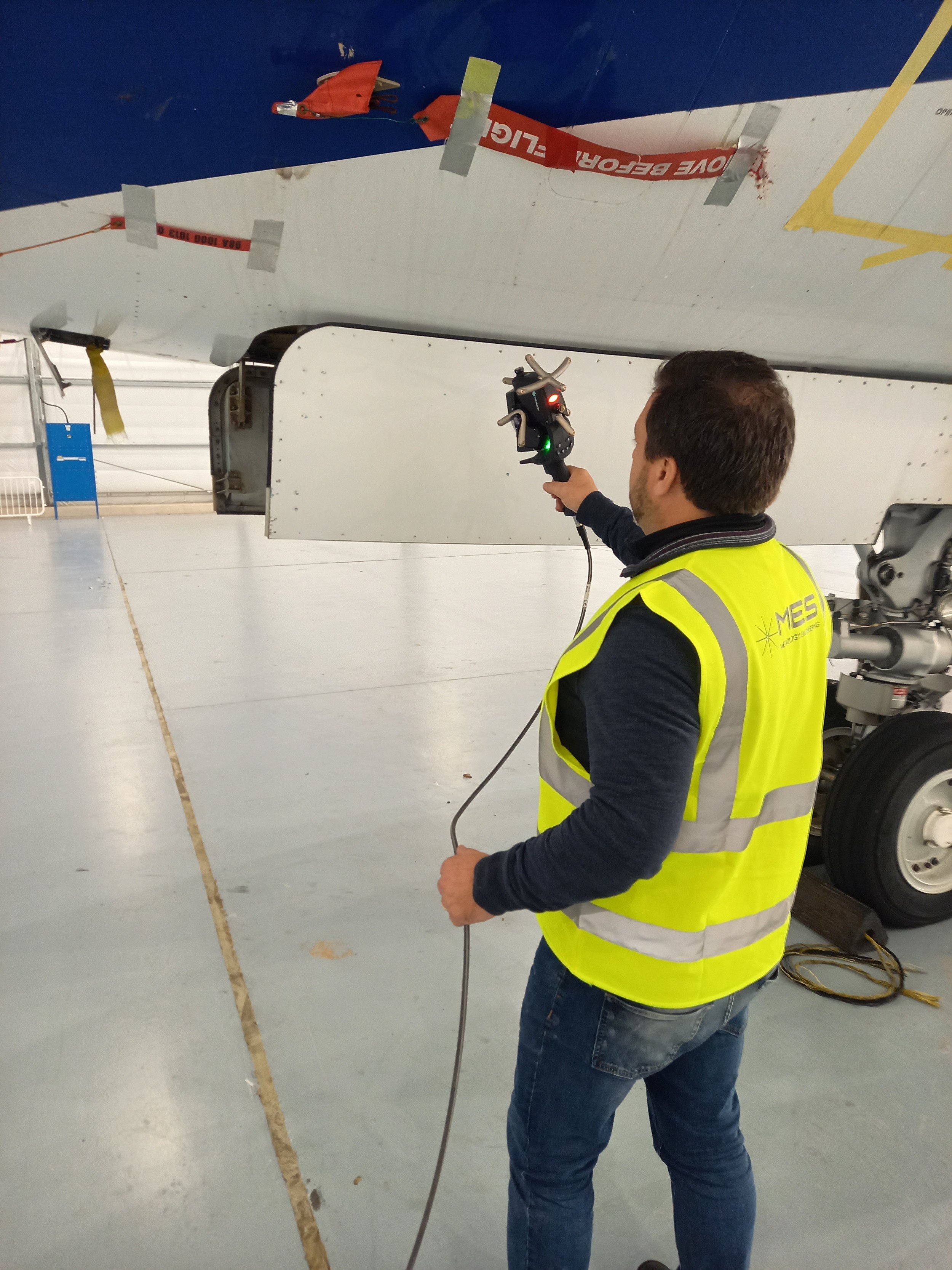 A man wearing a yellow safety vest and jeans is holding a device and working on an aircraft wing inside a hangar. The aircraft wing has a red safety warning and tape, with some tools and equipment nearby.