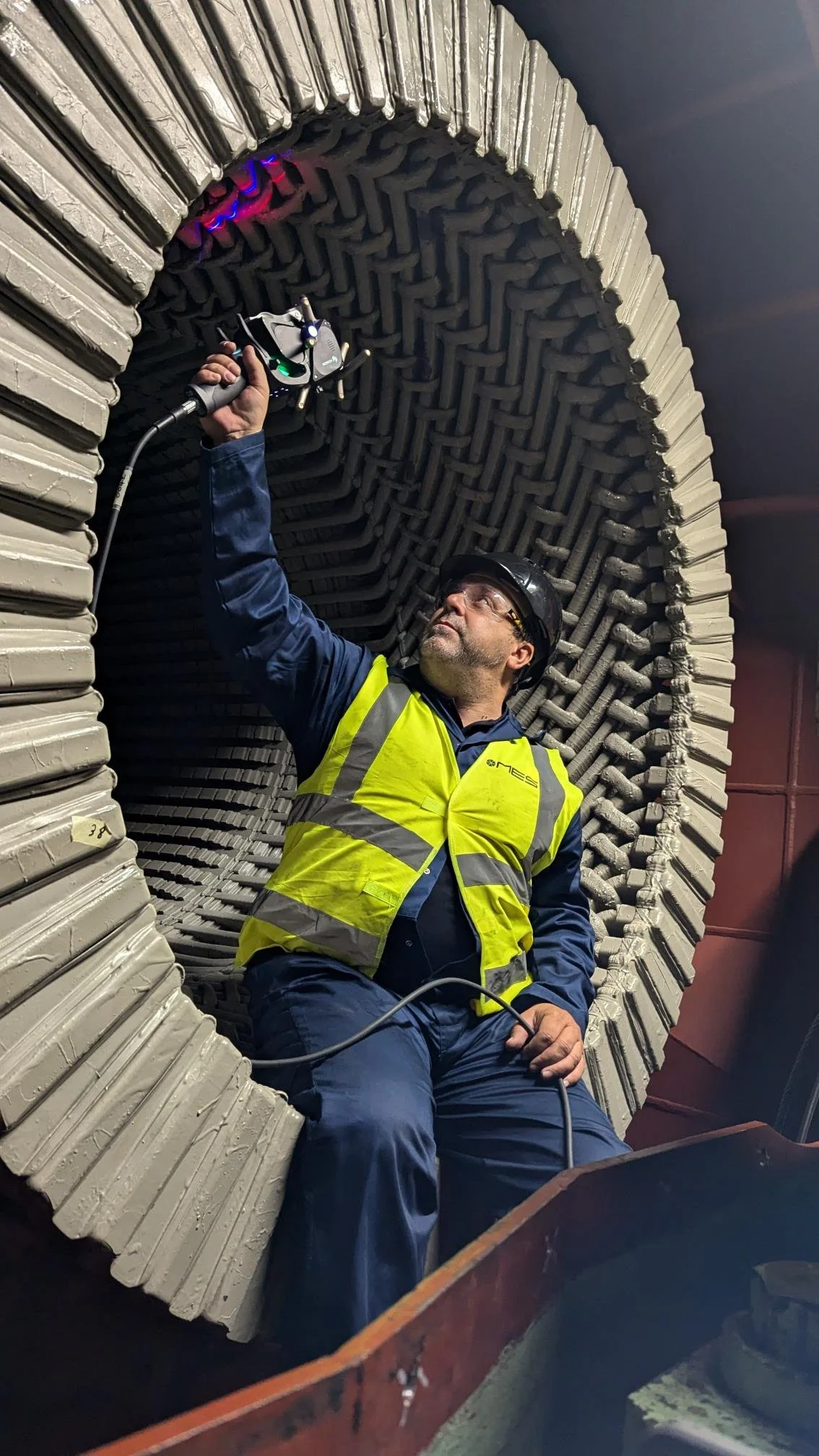 A man wearing safety glasses, a black helmet, and a yellow safety vest is sitting inside a large industrial tunnel made of metal pipes. He is holding and inspecting a piece of equipment or sensor attached to the tunnel's interior wall.