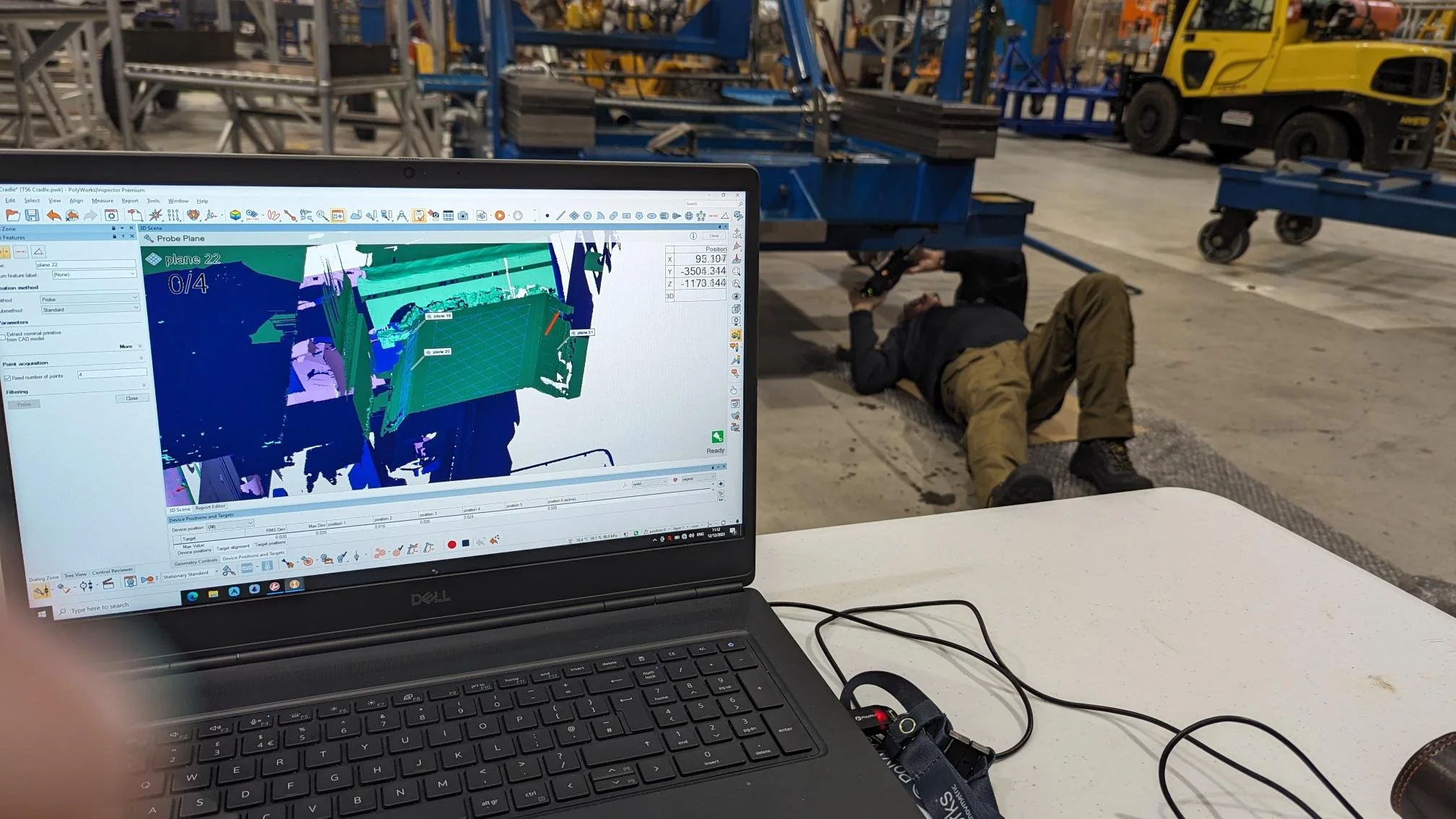 A laptop computer displays 3D modeling software with a colorful wireframe model on the screen. In the background, an MES worker is underneath a blue industrial structure in a warehouse or factory setting.