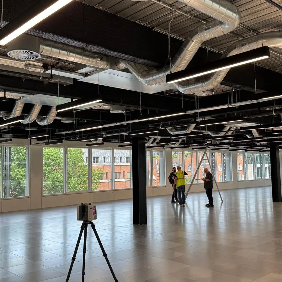 Interior of a modern office space with large windows, visible ductwork on the ceiling, and a group of construction workers discussing while installing or inspecting equipment.