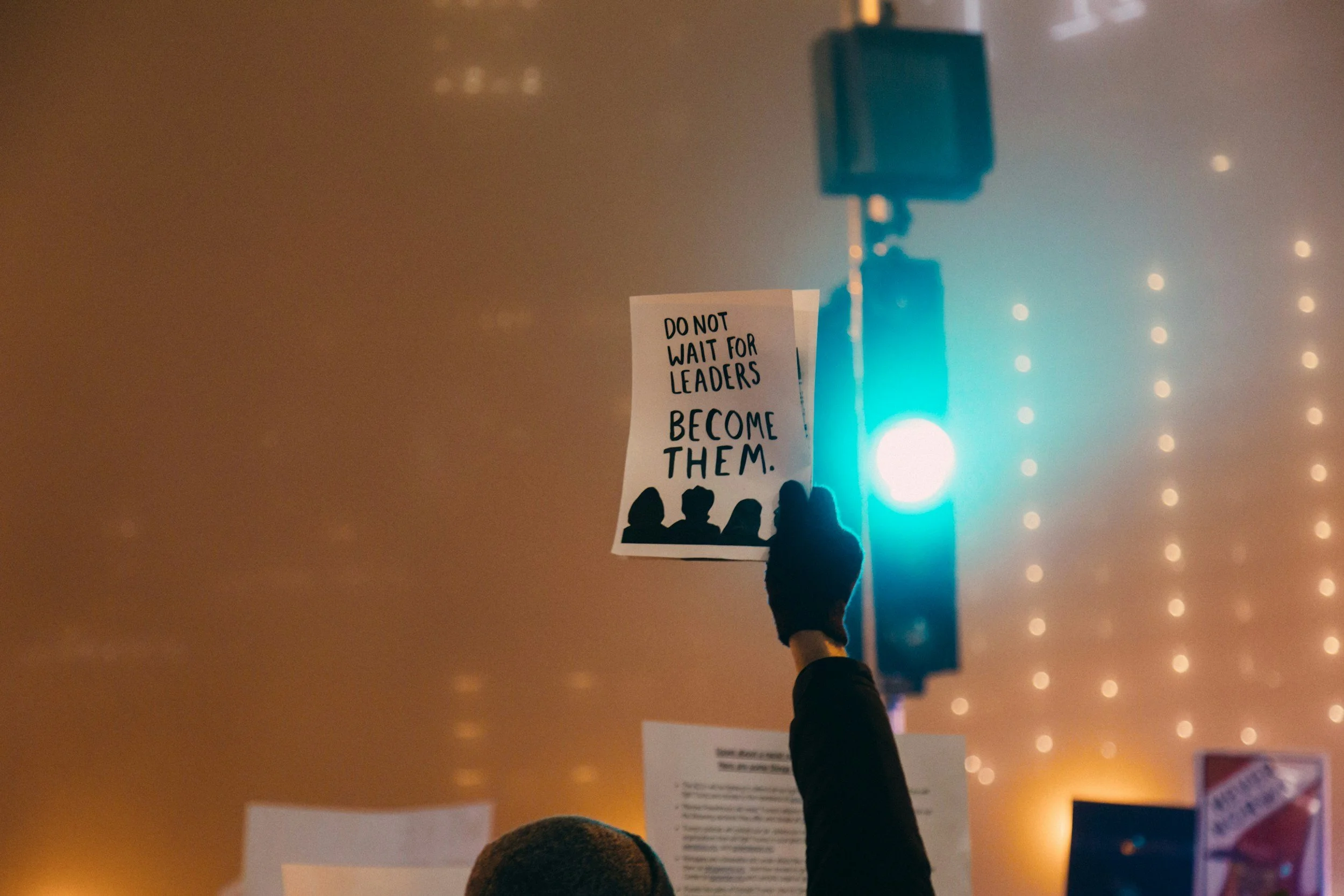 Eine Hand hält ein Plakat hoch, das den Text ‚DO NOT WAIT FOR LEADERS, BECOME THEM’ zeigt, bei einer Demonstration oder Versammlung bei Nacht mit Lichter im Hintergrund.