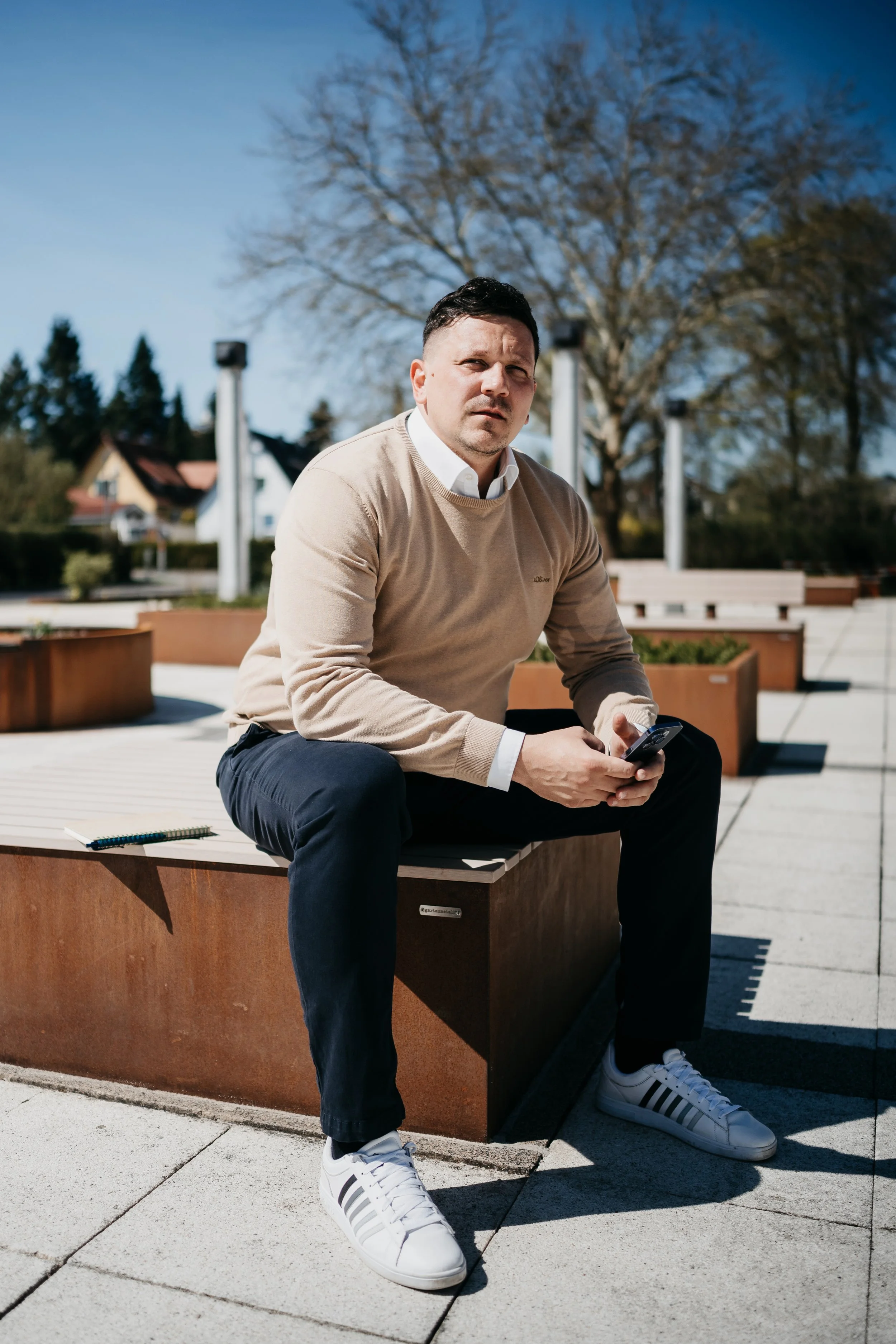 Ein Mann sitzt draußen auf einer Bank und schaut ernst in die Kamera, während er ein Smartphone hält.