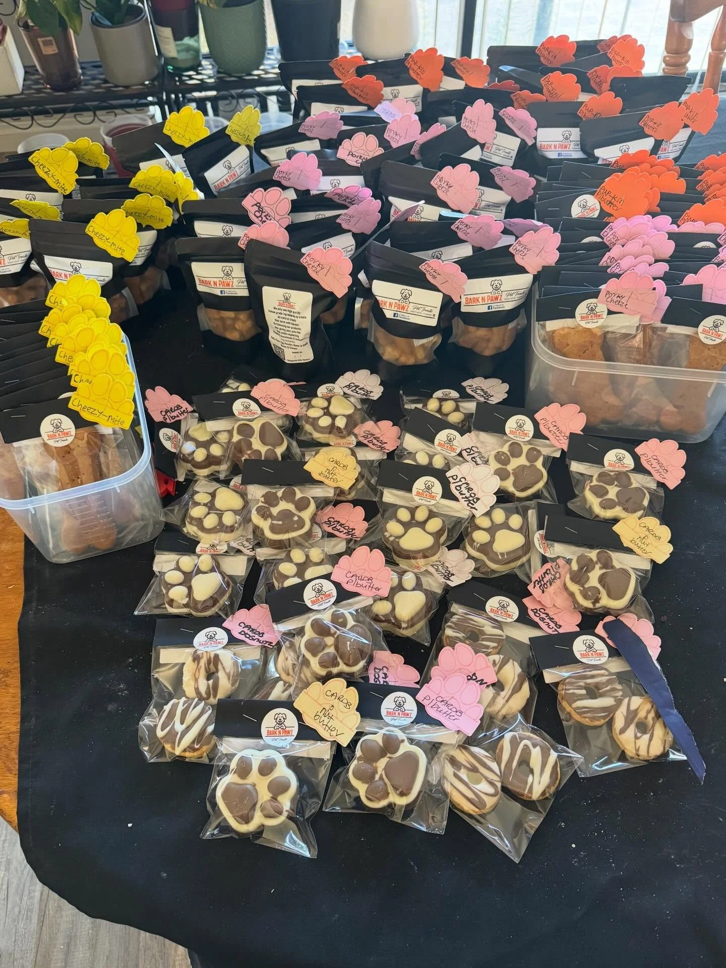 Assorted cookies in black, clear, and brown bags with dog-themed labels and pink, orange, white, and yellow tags on a black table. Some cookies are decorated with chocolate drizzle and paw prints.