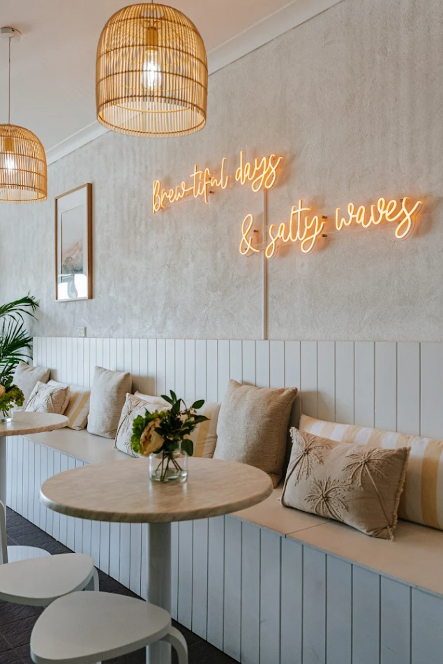 Cozy cafe with a bench seat lined with beige pillows, round tables with flower vases, soft lighting, and a neon sign on the wall reading 'breathtaking days & salty waves.'