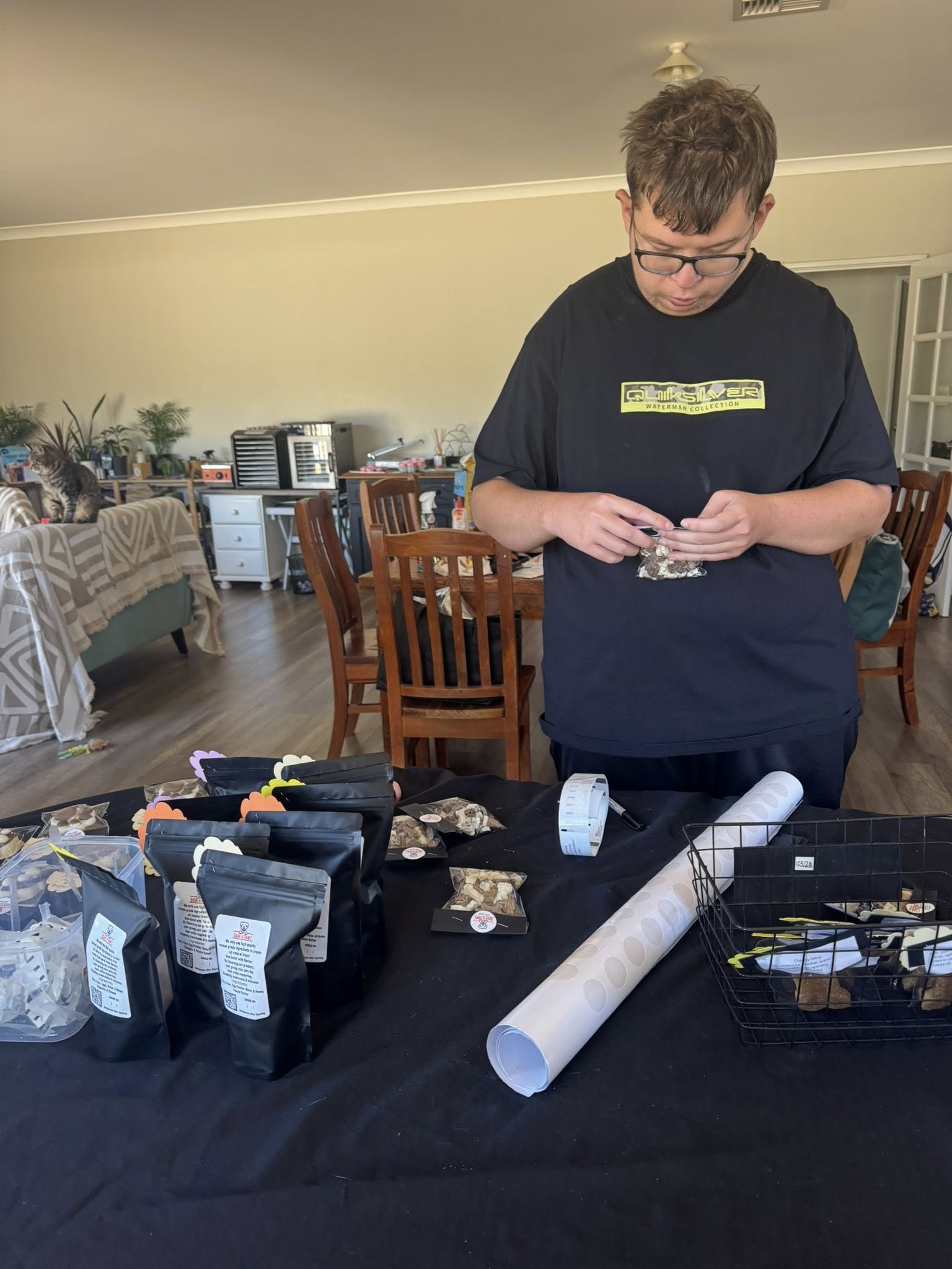 A man with glasses and short brown hair is standing at a table sorting small packages wrapped in brown paper, with several black bags and a roll of wrapping paper on the table; in the background, a living room with chairs, a couch, and various household items is visible.