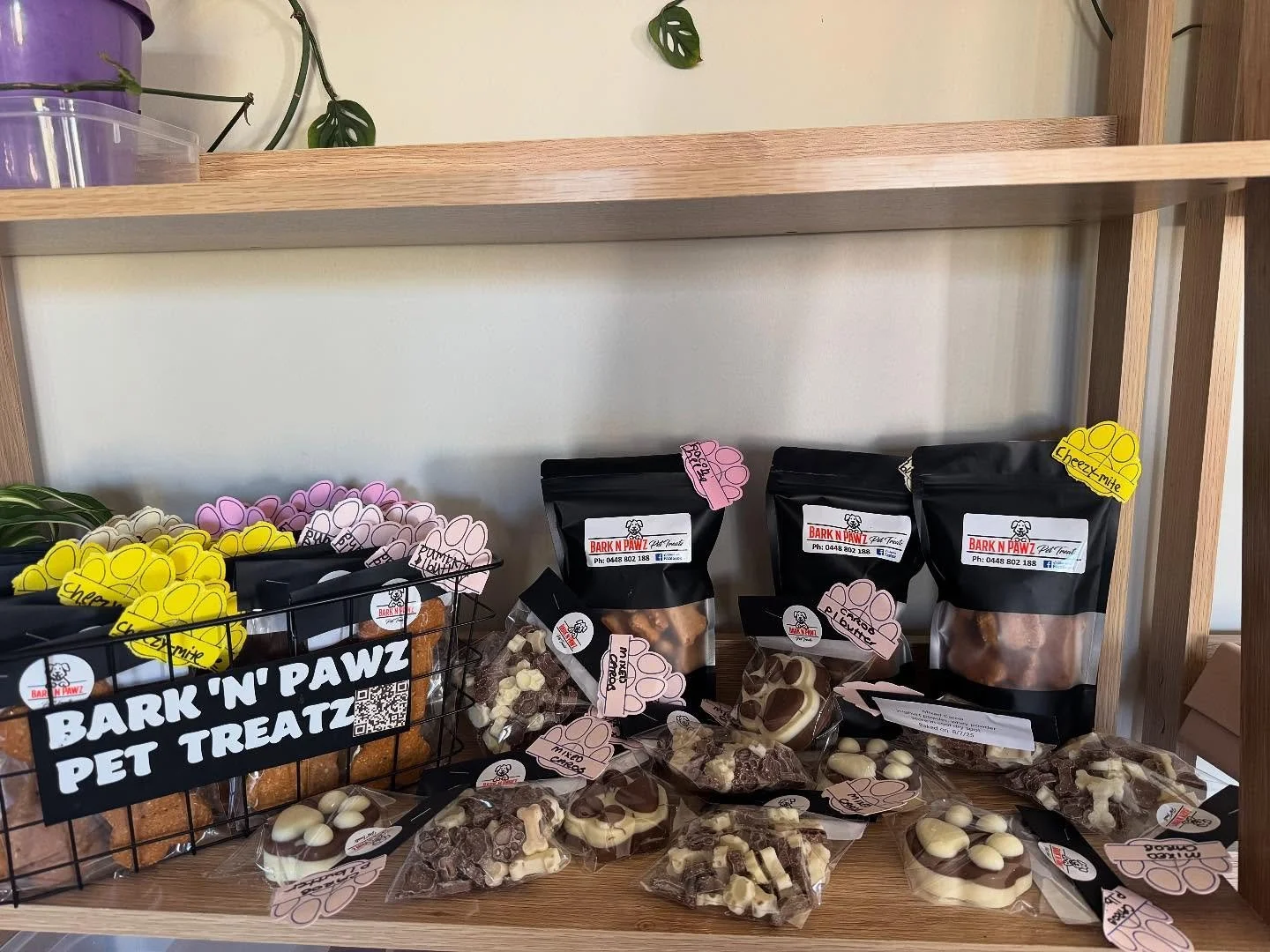 Display of various pet treats on wooden shelf, including packaged Bones, Chewy Mite, and assorted shaped treats in plastic bags and a wire basket labeled 'Bark 'N' Pawz Pet Treatz'.