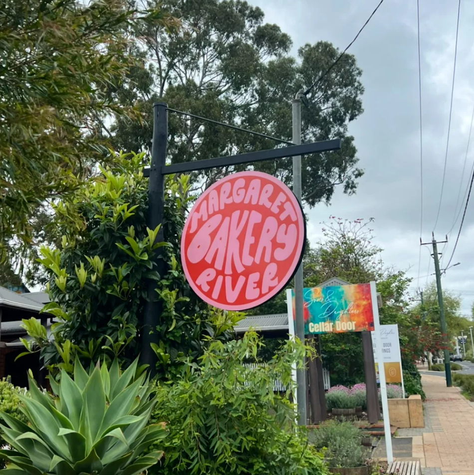 A round pink sign with white text reads 'Margaret Bakers Rive.' The sign hangs on a black metal post among green bushes and trees on a sidewalk in a small town.