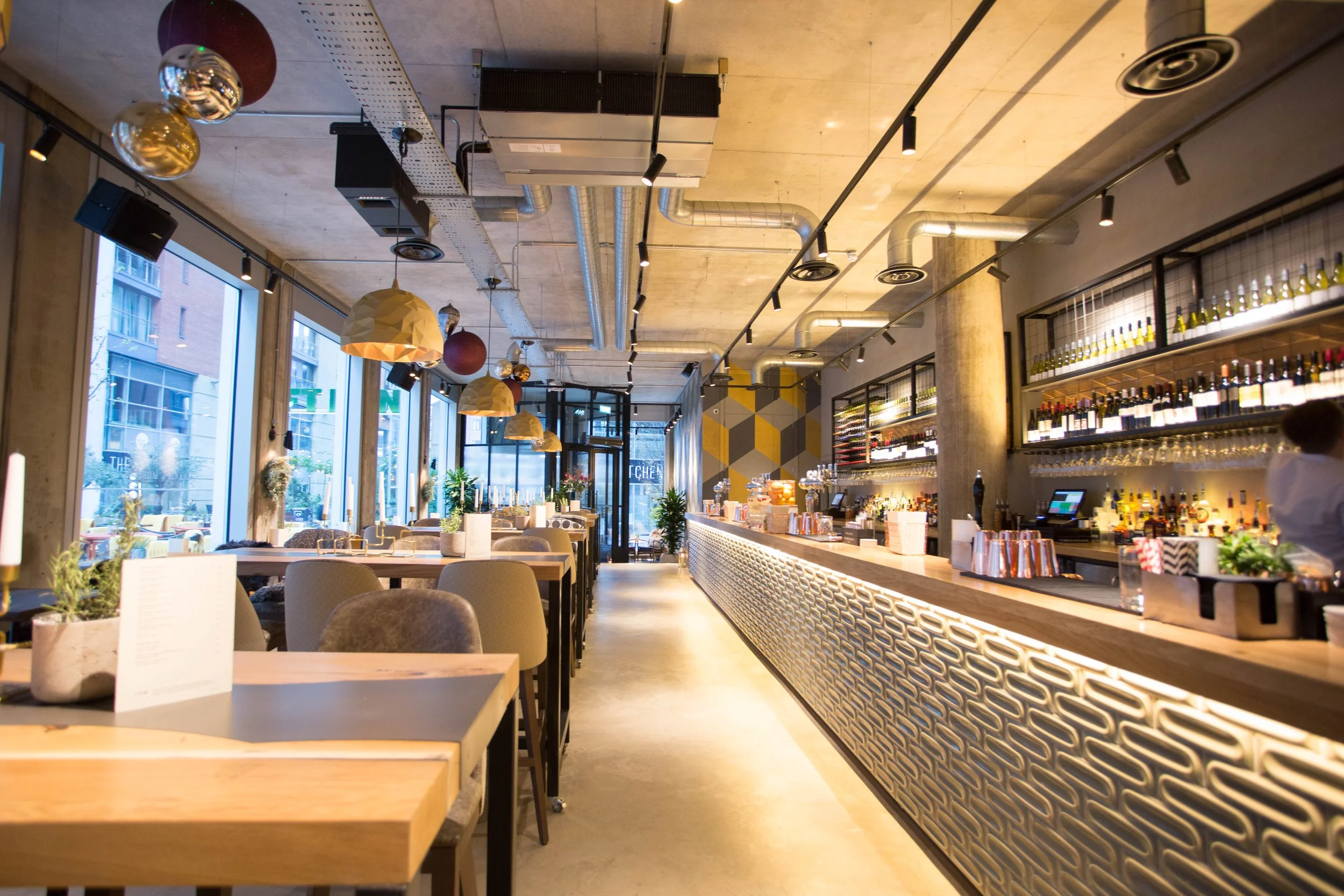 Interior of a modern restaurant with wooden tables, beige chairs, large windows, pendant and track lighting, shelves filled with wine bottles, and a bar counter with various bottles and glasses.