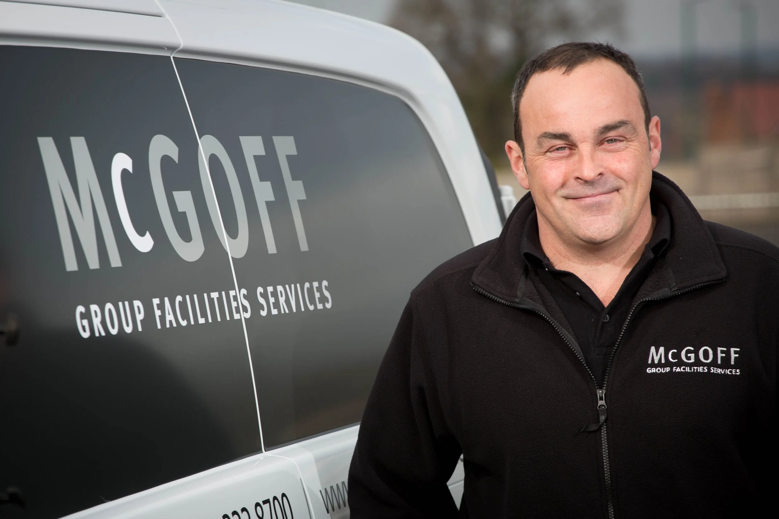 A smiling man wearing a black jacket with the text "McGOFF GROUP FACILITIES SERVICES" standing next to a vehicle with the same company name and logo on the side.