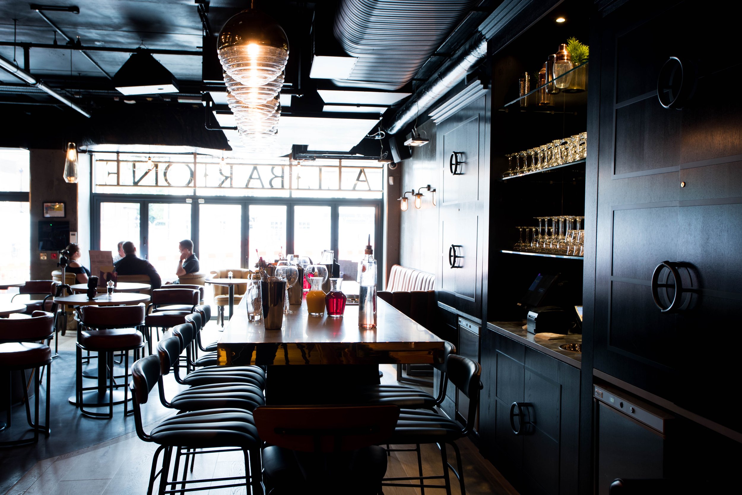 A modern restaurant interior with a long wooden bar counter, arranged with glasses and bottles, and seating for customers. Large windows allow natural light to illuminate the space, which contains several tables and chairs. The decor includes dark wa