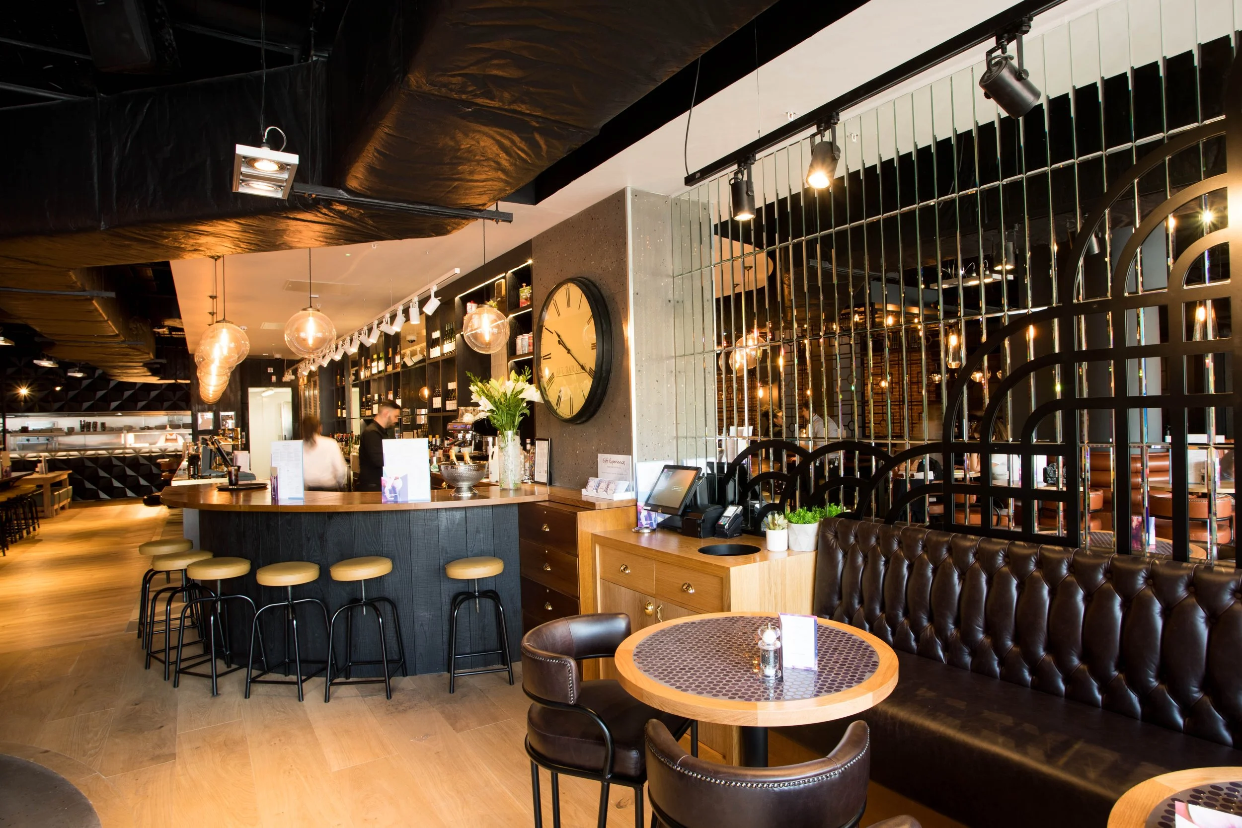 Interior of a modern cafe or restaurant with a counter, bar stools, tables, and chairs, decorated with hanging lights, a clock, and plants.