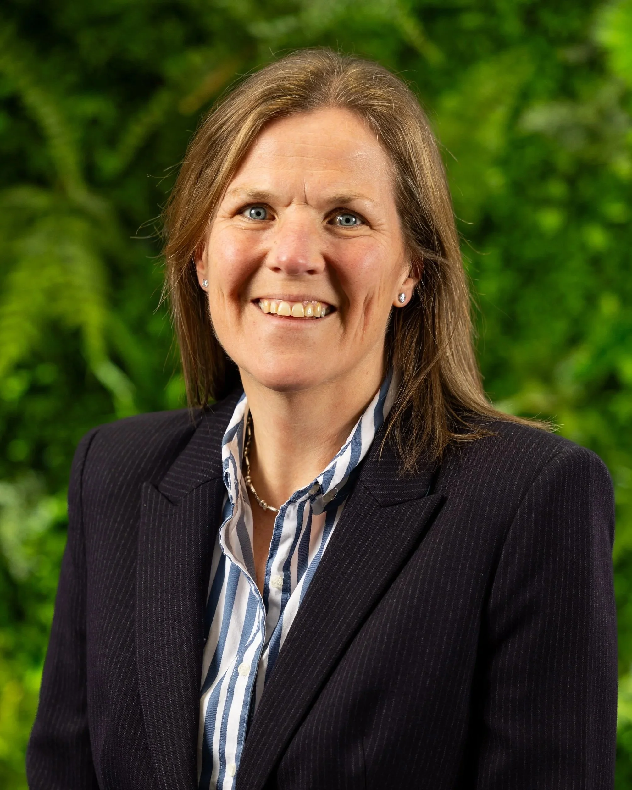 A smiling woman with blue eyes and brown hair wearing a striped shirt and black blazer, standing outdoors with green foliage in the background.