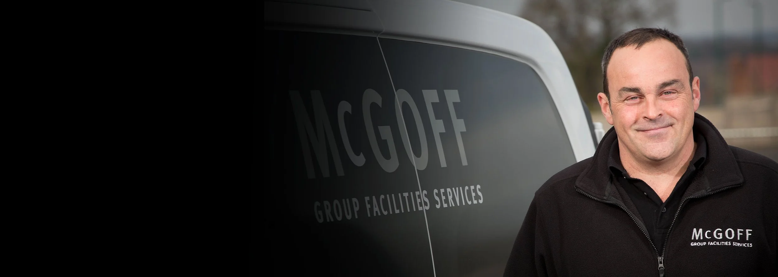 A man smiling outdoors standing in front of a vehicle with the words 'MCGOFF GROUP FACILITIES SERVICES' written on it. The man is wearing a black jacket with the same company name embroidered on it.