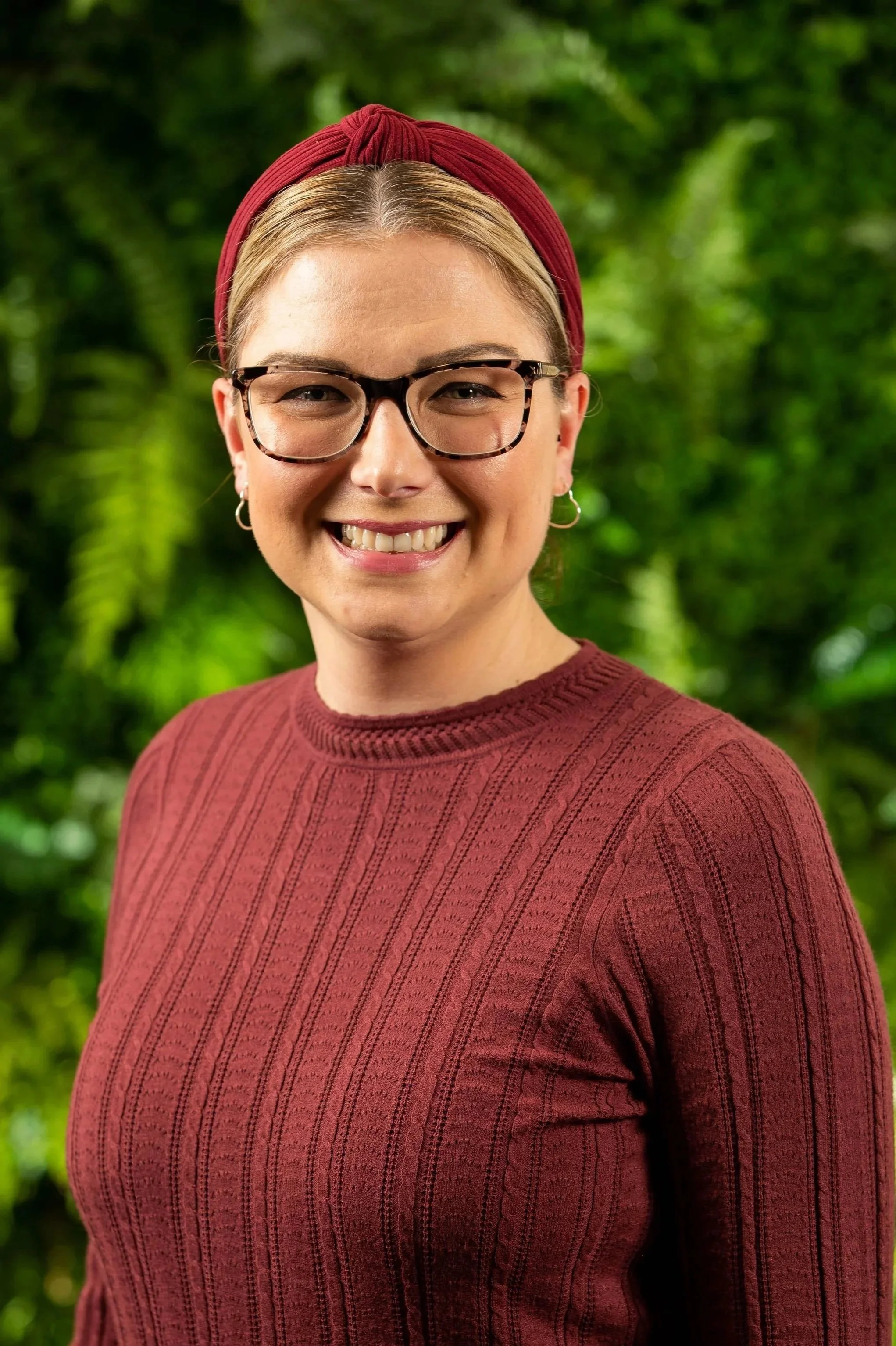A woman smiling outdoors with a green leafy background, wearing glasses, a red headband, and a red sweater.
