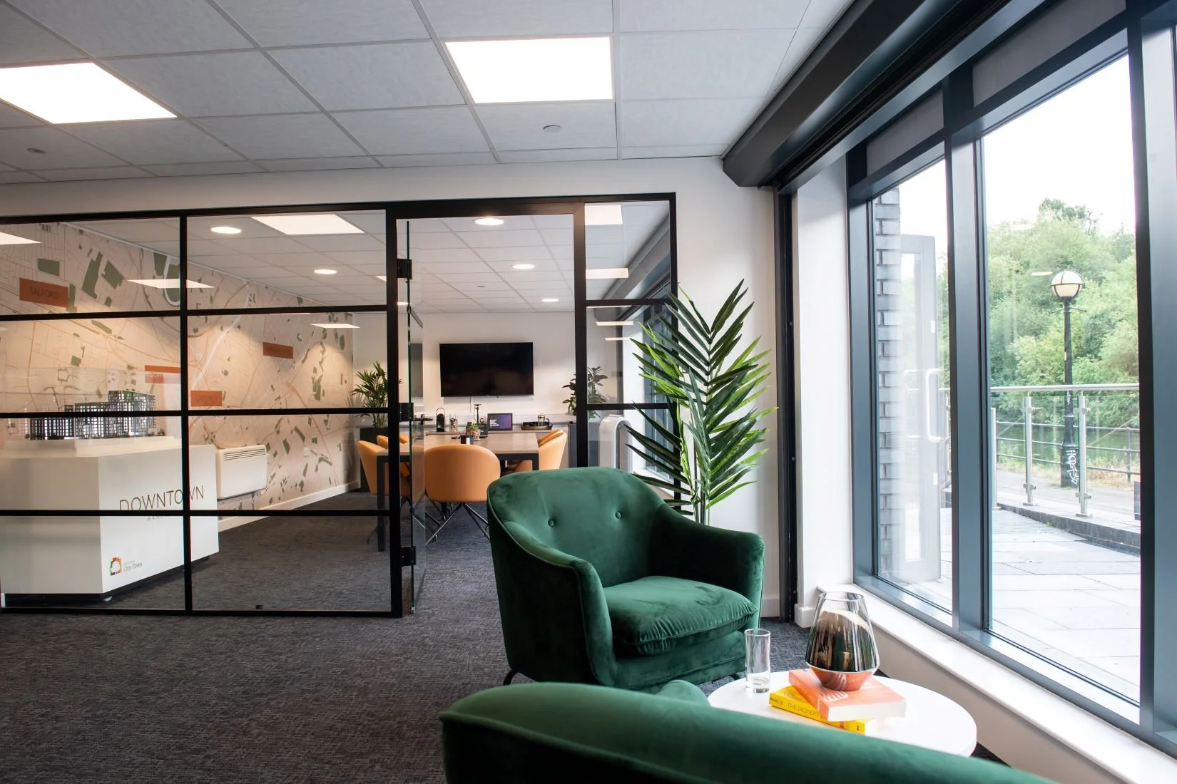 Modern office space with large windows, a green armchair, a glass of water, books, and a view of trees outside.