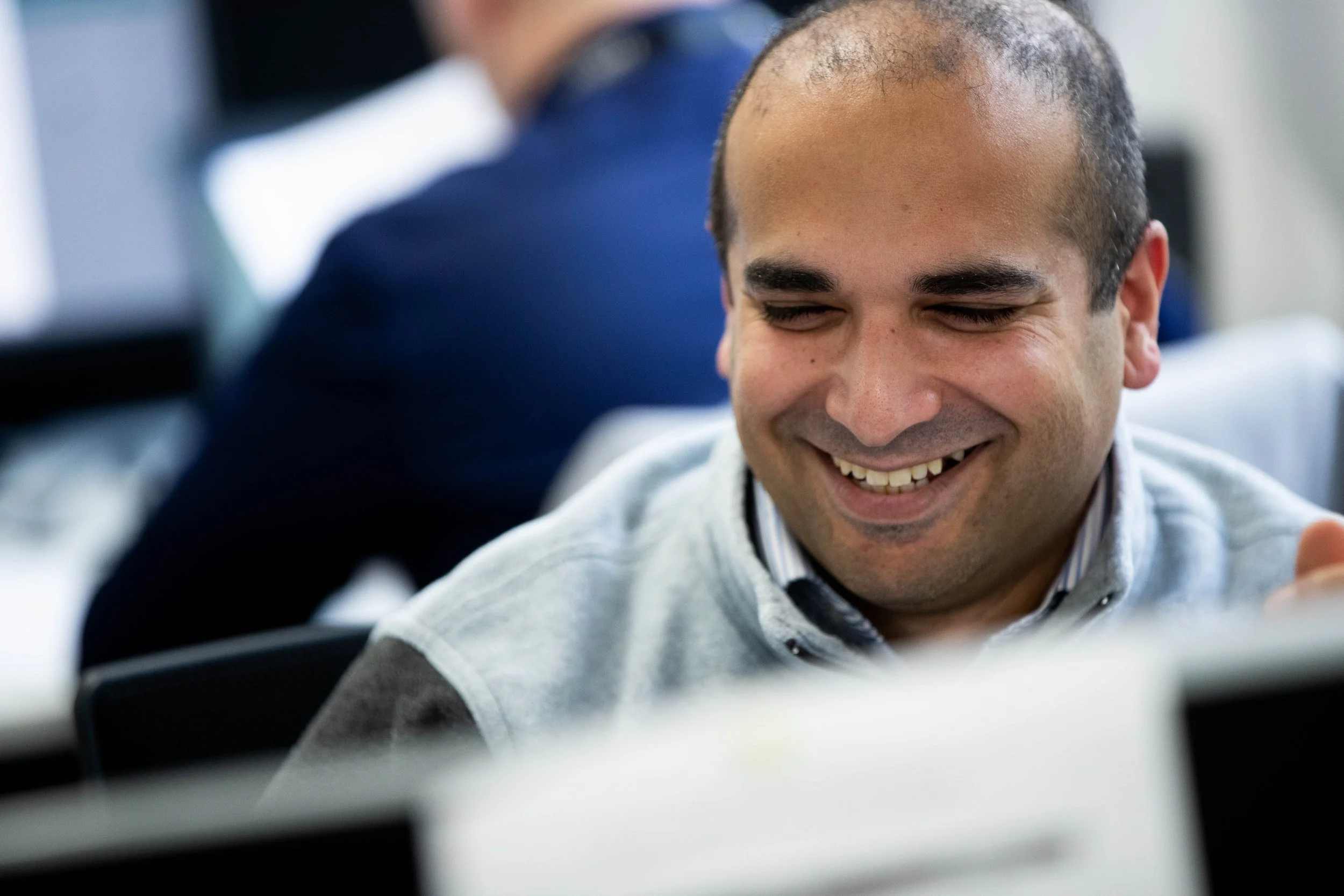 A man smiling with his eyes closed, wearing a light gray shirt, in an indoor setting with blurred people in the background.