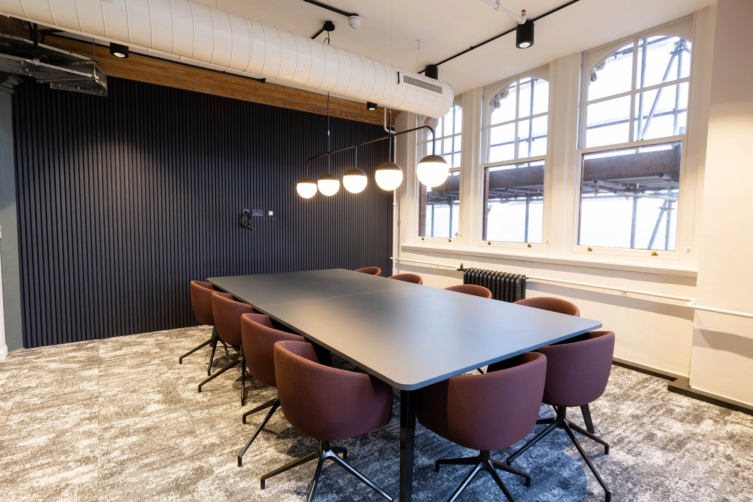 Modern conference room with a large rectangular table surrounded by pinkish purple chairs, black wall paneling, and large windows with a cityscape view, illuminated by hanging globe pendant lights.